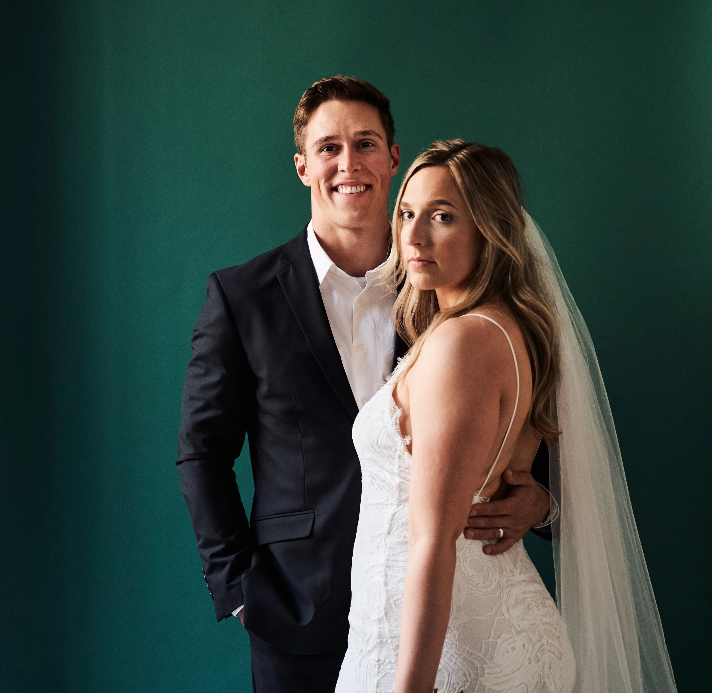 A young couple in wedding attire standing back to back against a green backdrop. The man is dressed in a black suit with a white shirt, smiling, facing forward. The woman is wearing a white lace dress with spaghetti straps, with her face partly turne