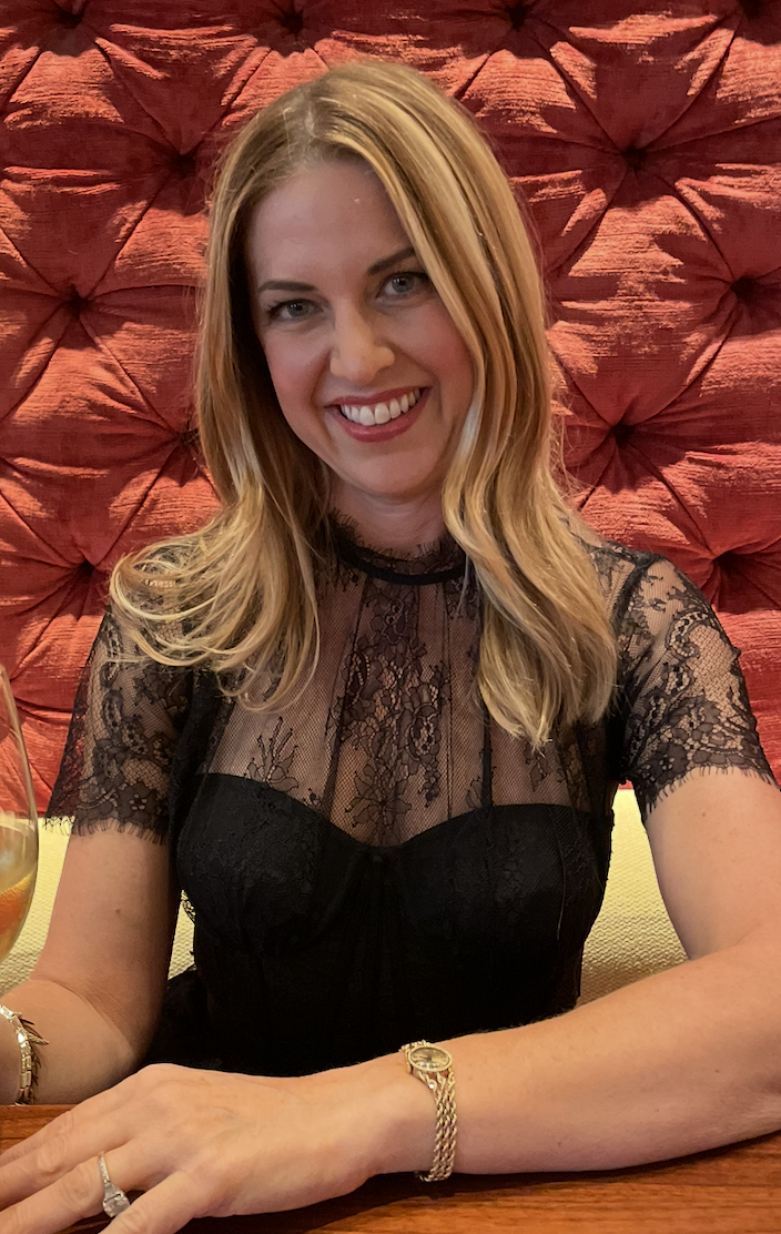 A woman with blond hair smiling, wearing a black lace top, sitting at a table in front of a red tufted background.