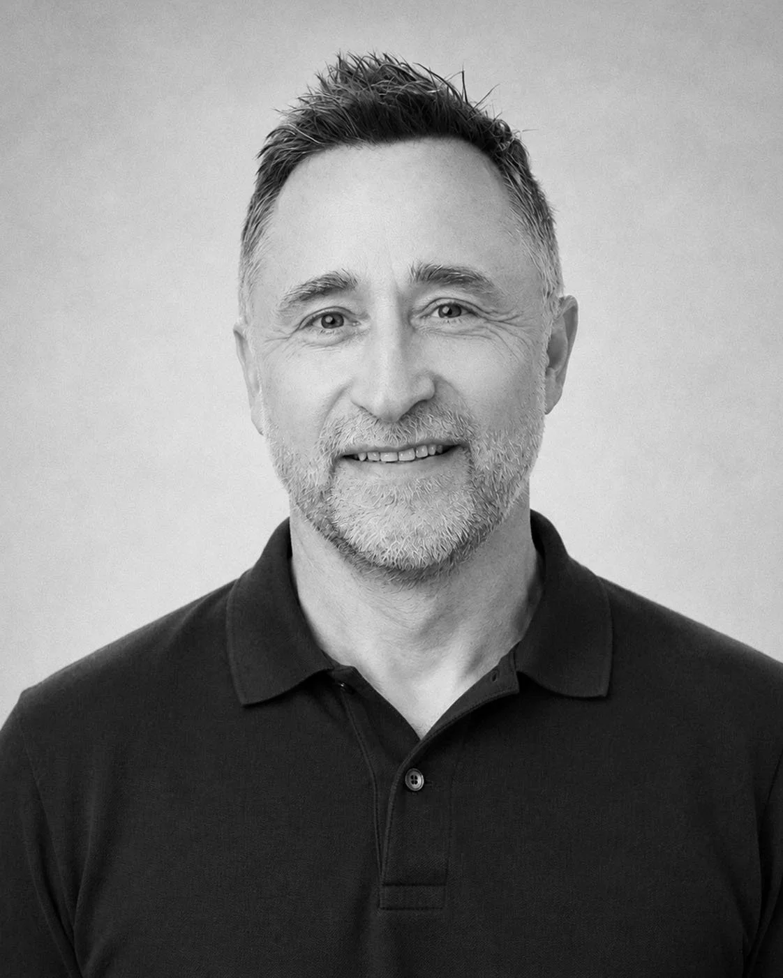 Black and white portrait of a middle-aged man with short hair and a beard, smiling gently, wearing a collared shirt.