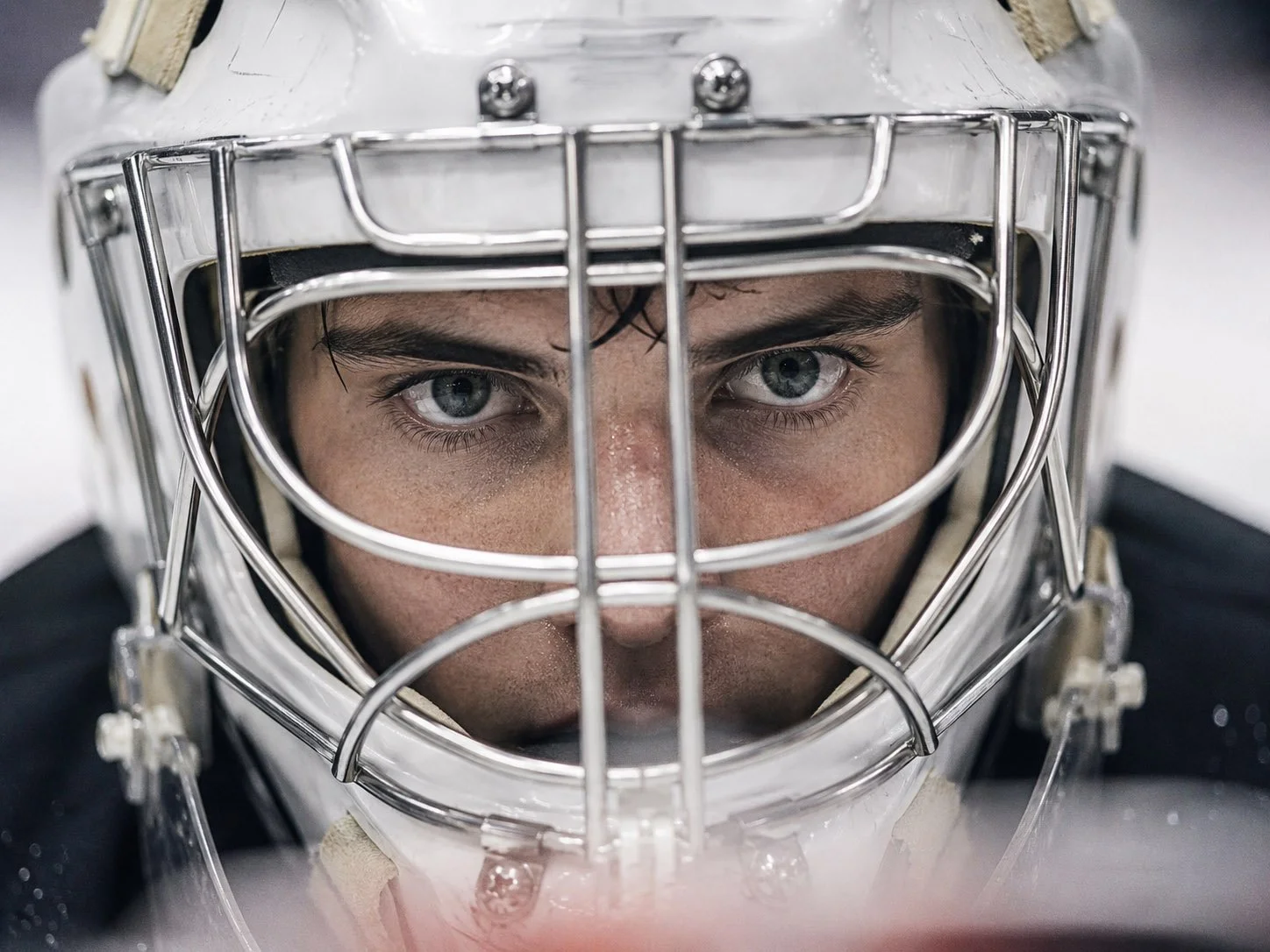 Close-up of a male ice hockey goalie with intense blue eyes, wearing a metal cage mask, looking straight ahead.