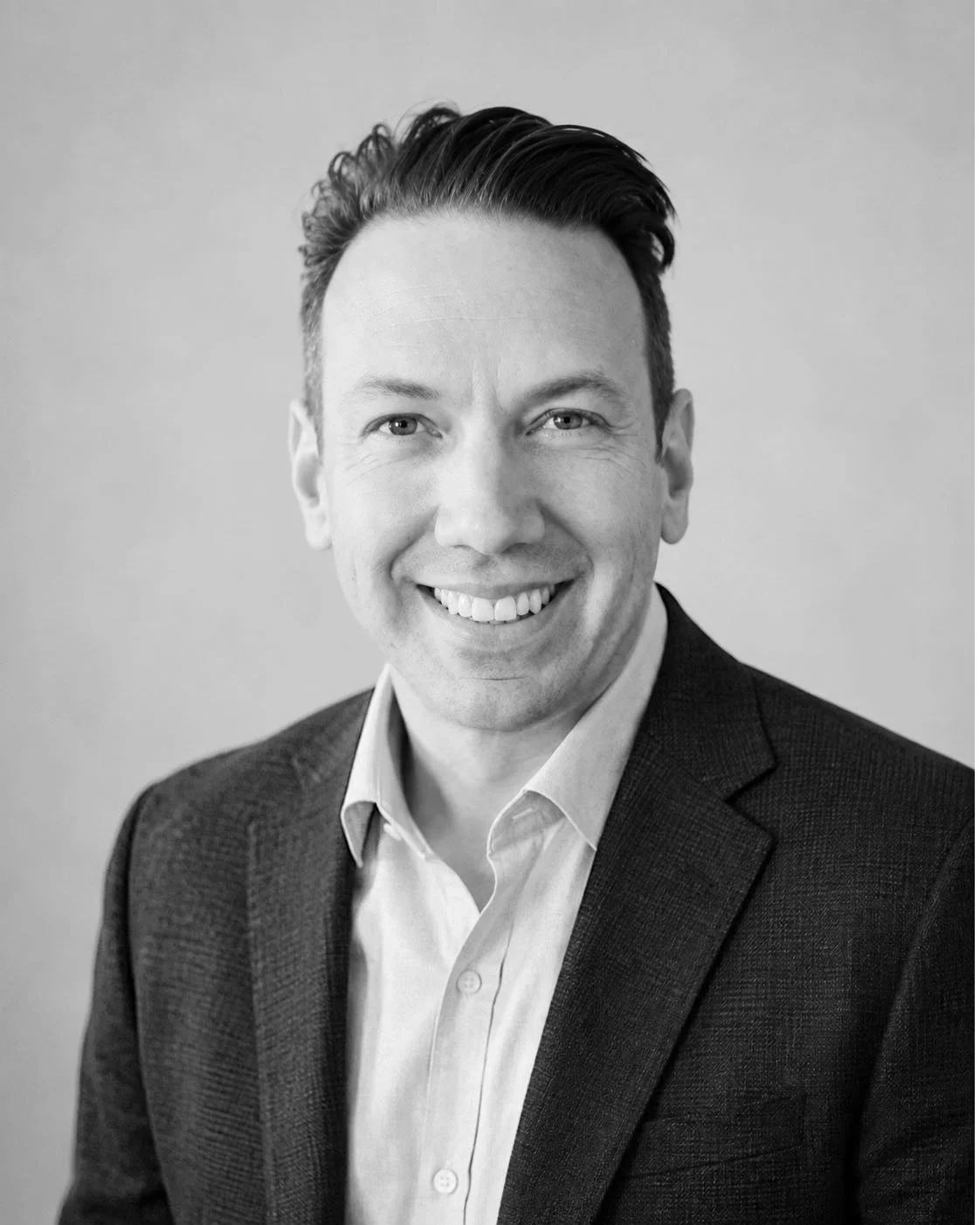 Black and white headshot of a smiling man in business attire, wearing a suit jacket and shirt, against a plain background.