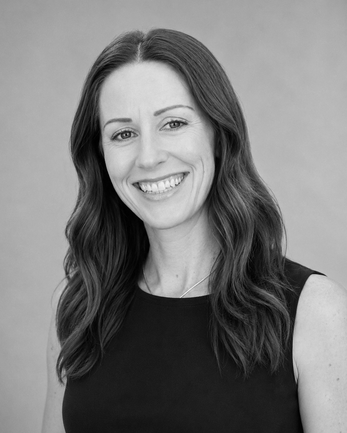 A black and white headshot of a woman with long wavy hair, smiling, wearing a sleeveless black top and a thin necklace, against a plain background.
