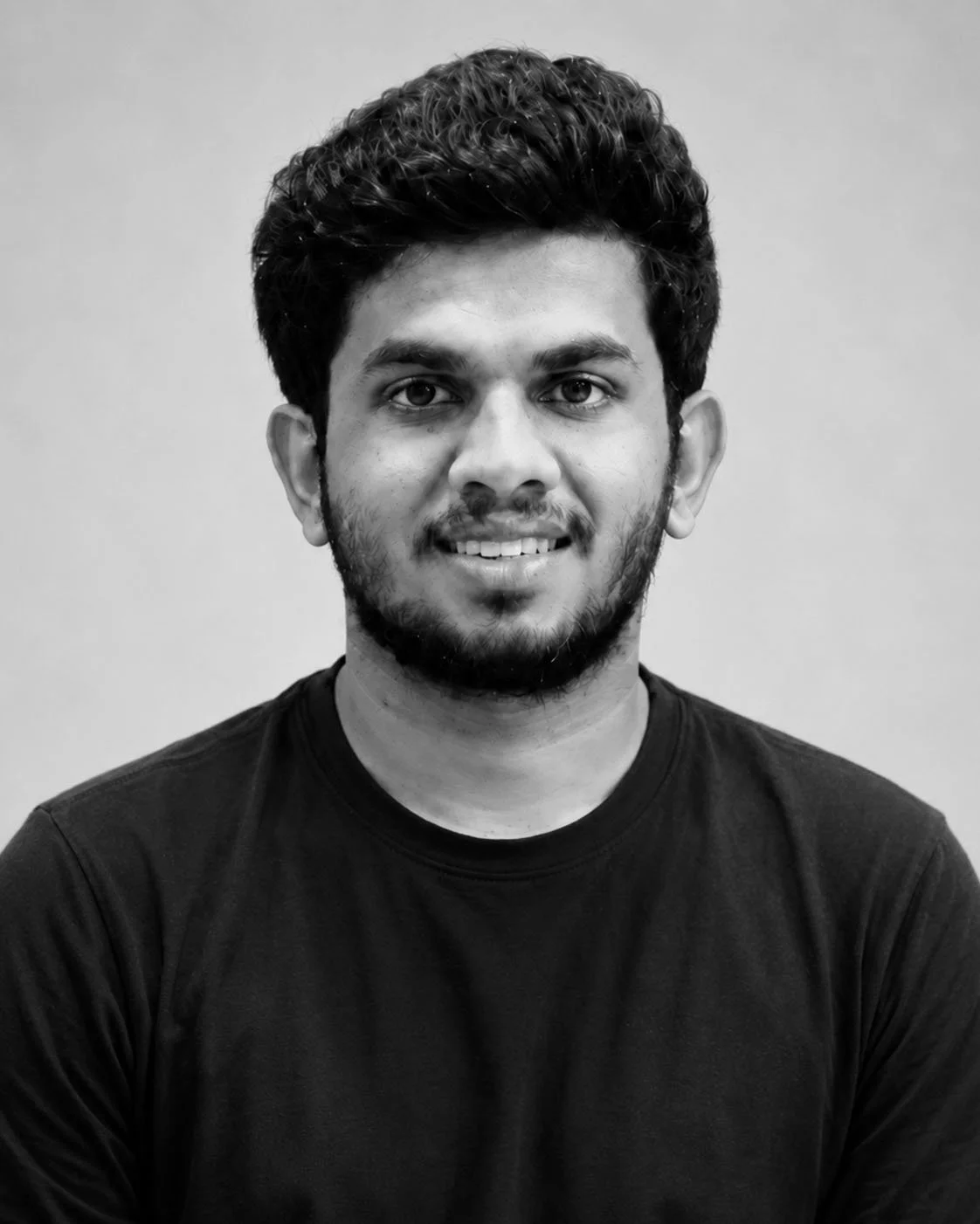 Black and white portrait of a young man with dark, curly hair, a beard, and mustache, wearing a dark t-shirt, smiling slightly, against a plain background.