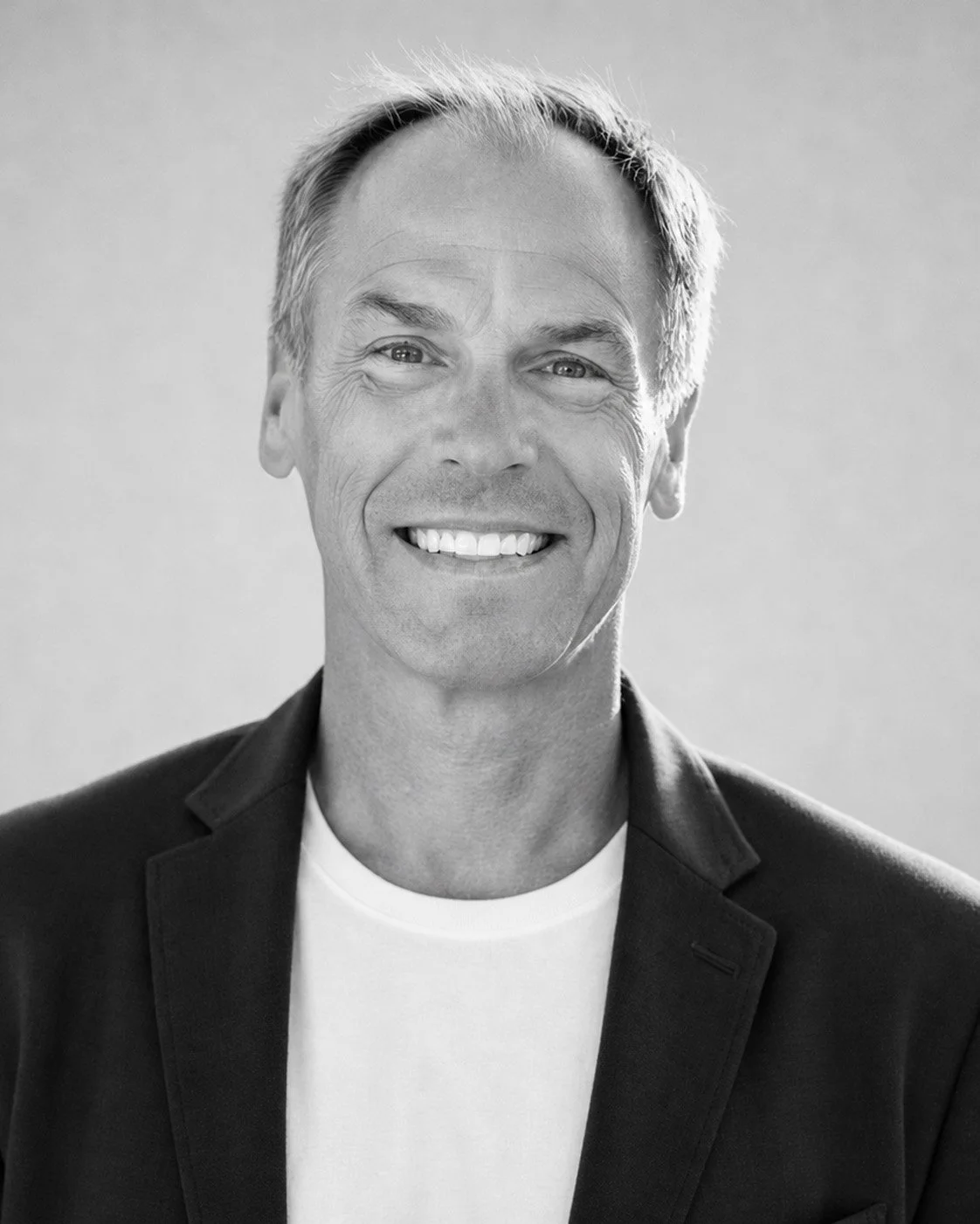 Black and white portrait of a smiling man with light-colored hair, wearing a dark blazer over a white shirt.