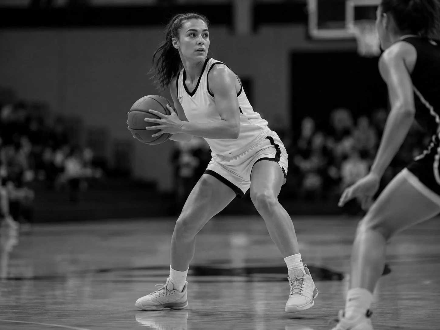 Black and white photograph of a female basketball player in a white uniform holding a basketball, possibly preparing to make a move, on an indoor court. Another player is partially visible to her right.