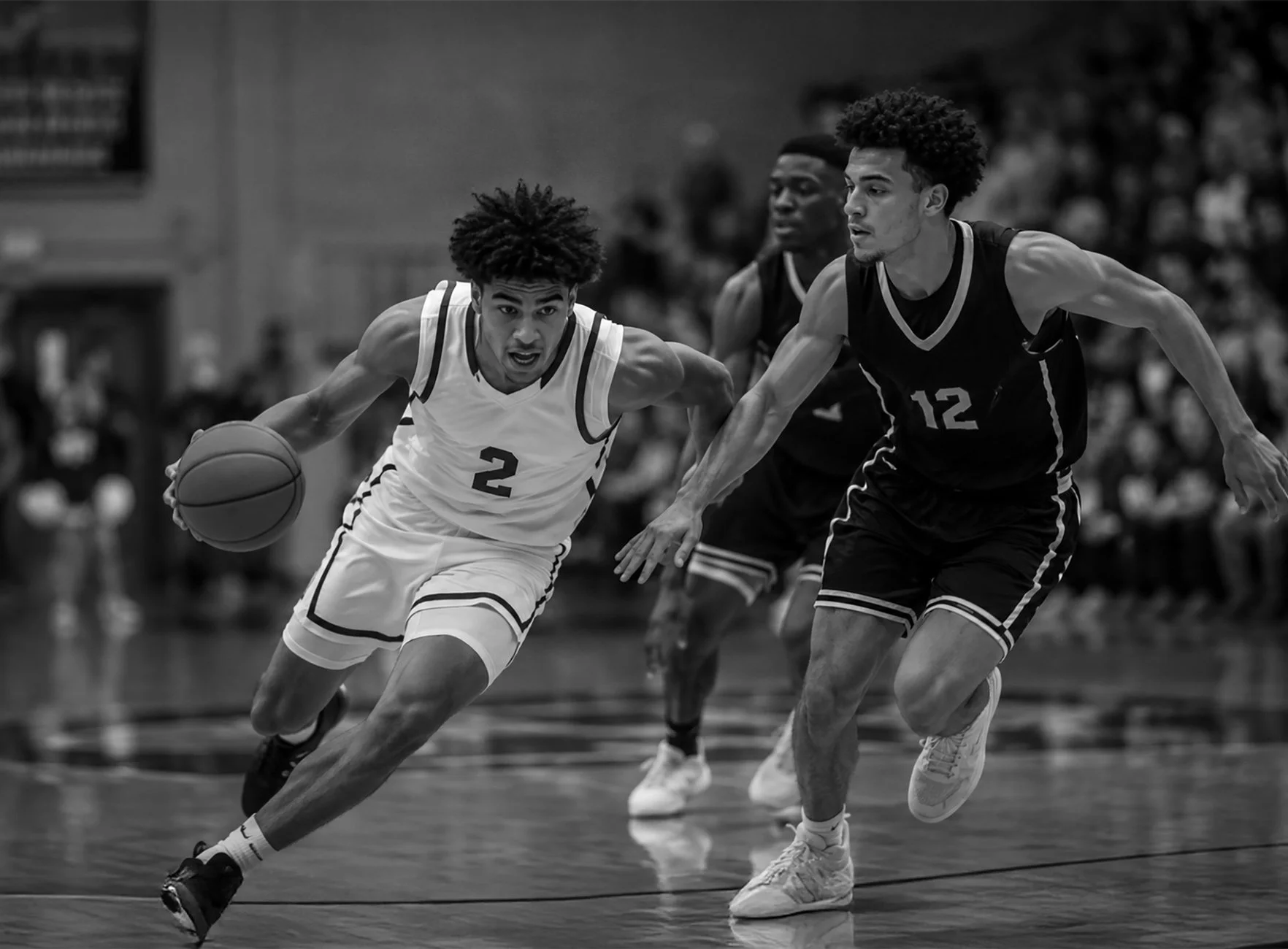 Black and white action shot of a basketball game showing two players racing for the ball on the court, with a crowd in the background.