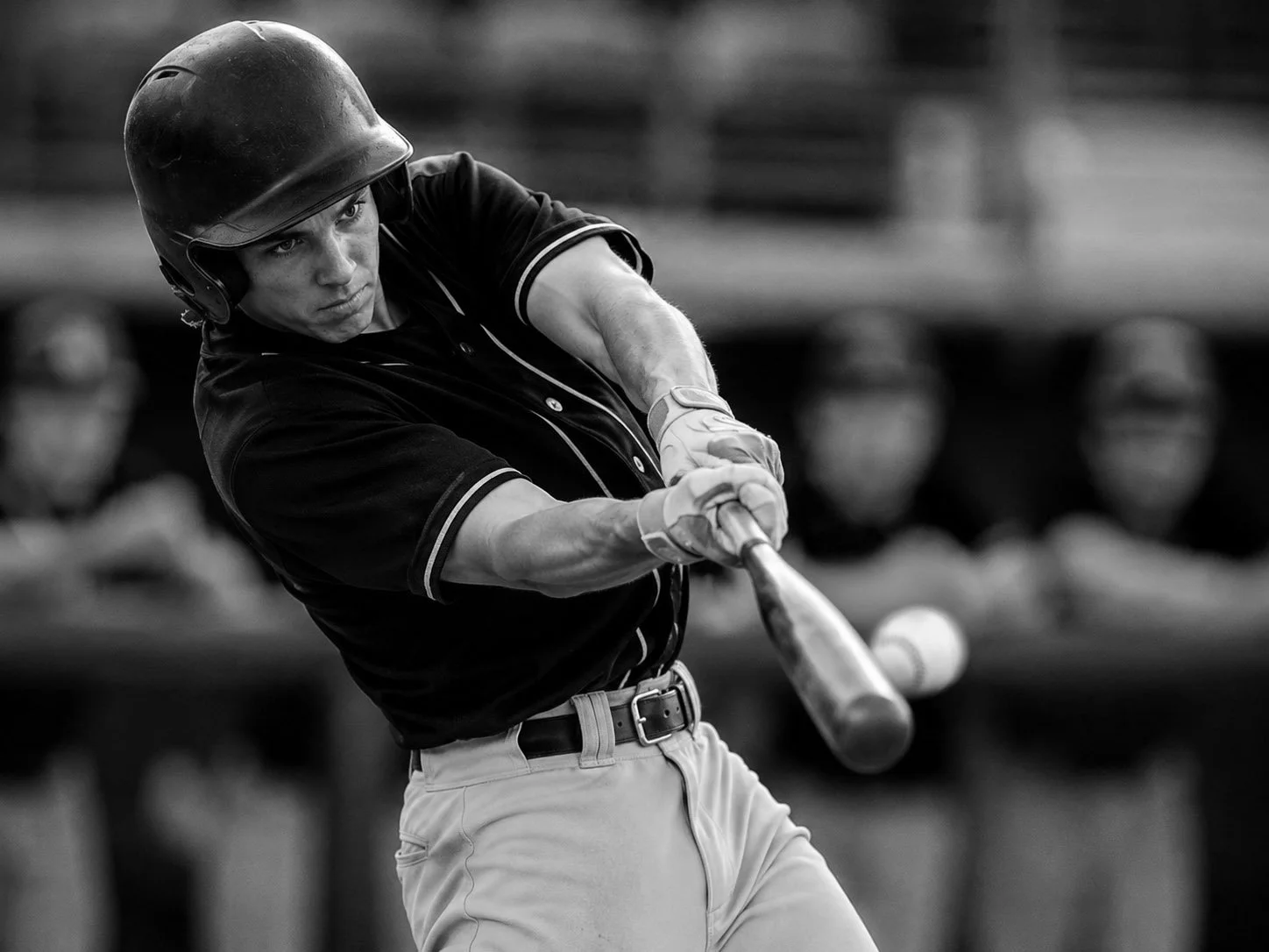 Black and white photo of a young man playing baseball, swinging a bat. He is wearing a helmet and a dark shirt with light-colored pants.