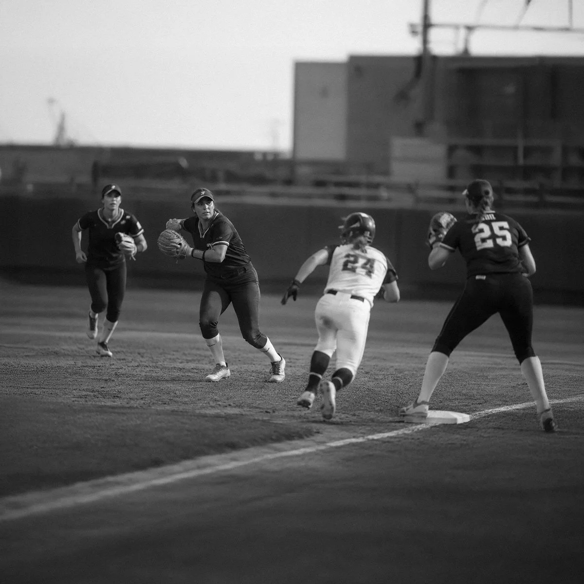 Black and white photo of a women's softball game, showing players running, catching, and sliding on the field during a game at dusk or night.