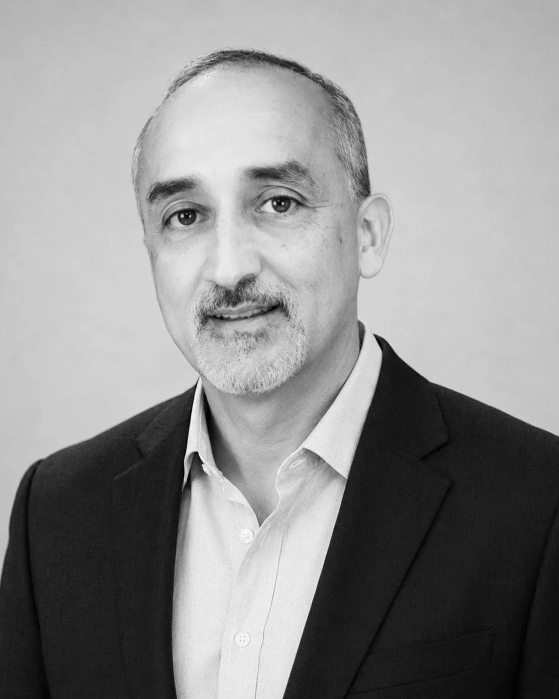 Black and white professional headshot of a middle-aged man wearing a suit and collared shirt, smiling slightly, with short hair and a goatee.