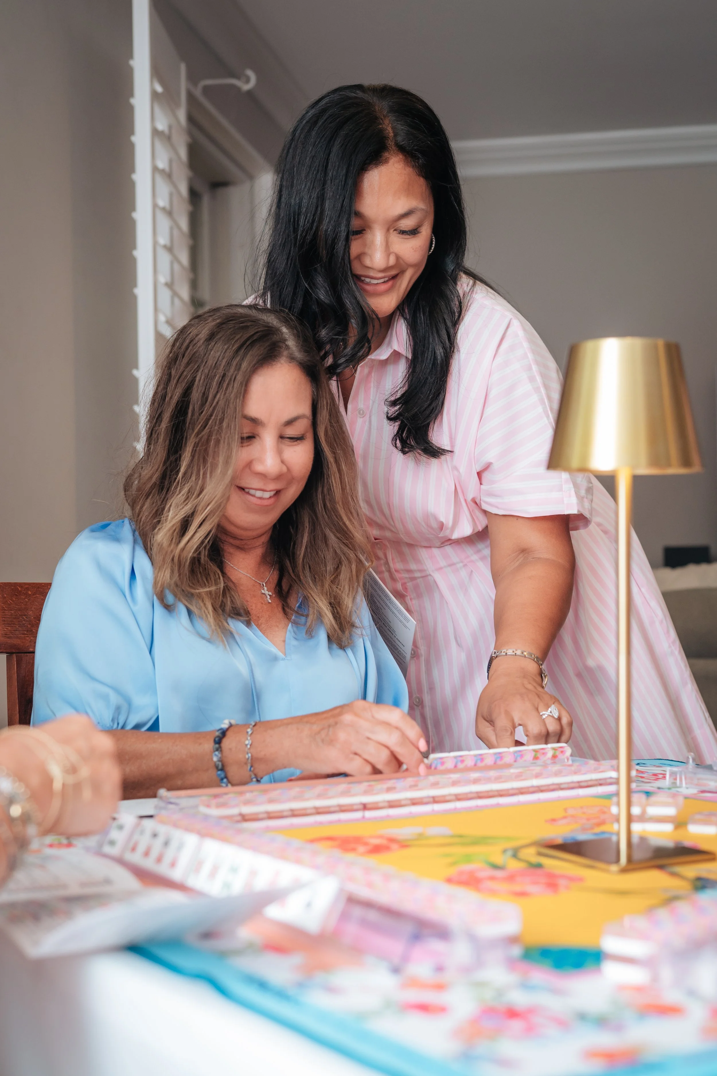 Two women, one standing and one sitting at a table, engaging with papers and games, in a cozy indoor setting with a gold table lamp.