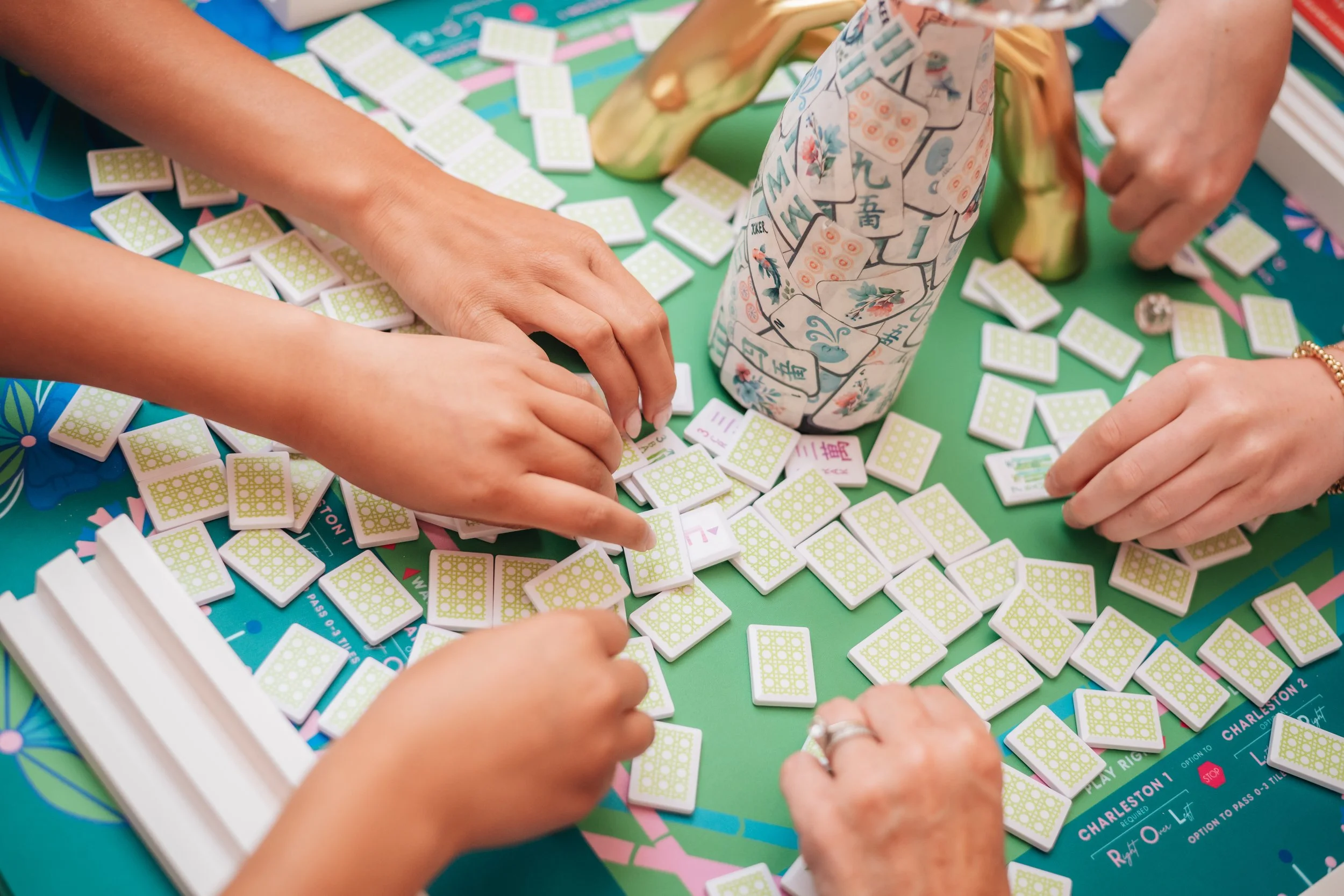 People playing Mahjong at a colorful table with green tiles and a paper-wrapped bottle.