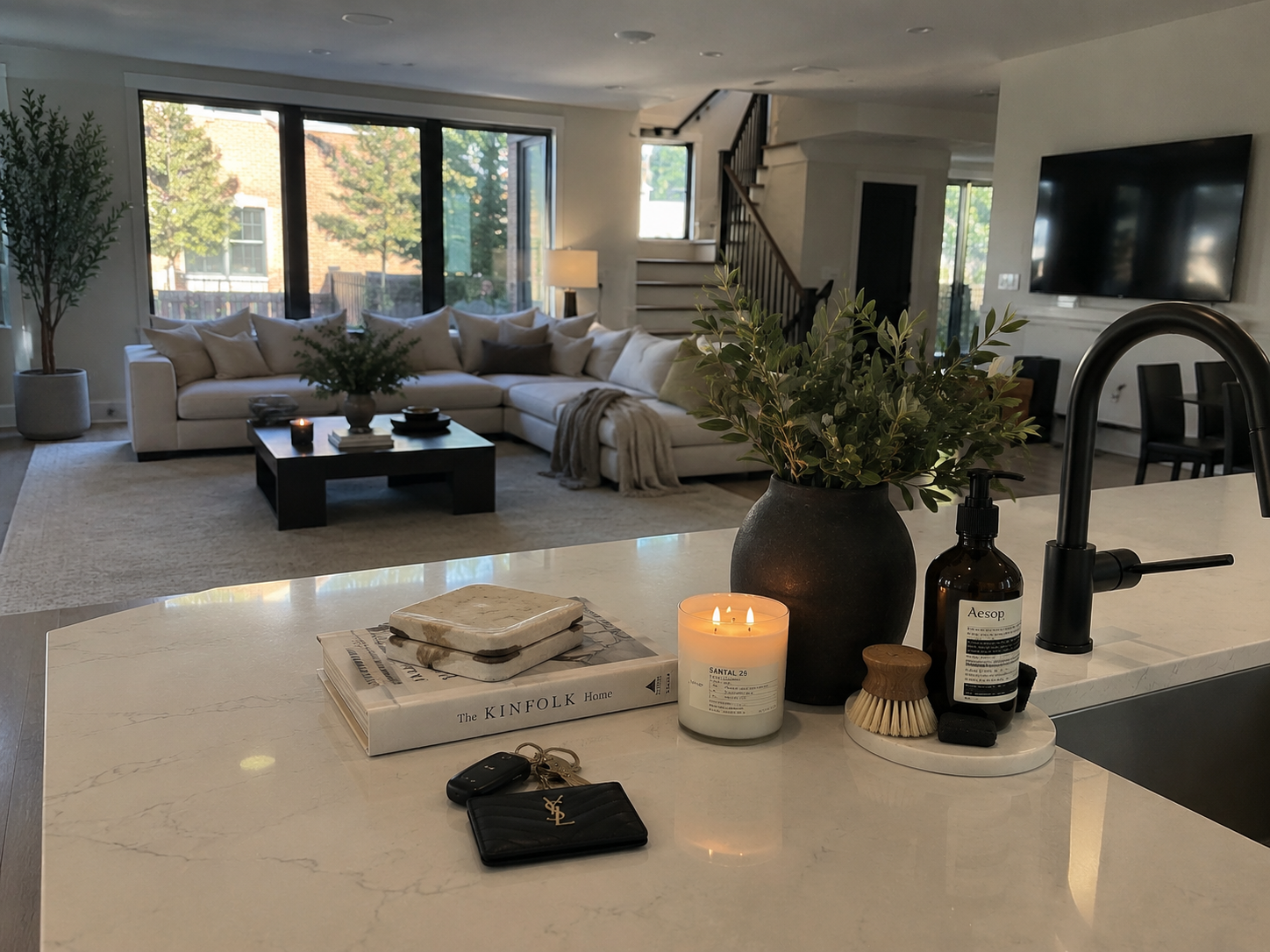 View of a modern living room seen from a kitchen counter. The living room features a white sectional sofa with multiple pillows, a black coffee table with a vase of greenery, large windows with trees outside, and a TV mounted on the wall. The kitchen counter has a candle, a black vase with greenery, a bottle of hand soap, a brush, and a set of keys with a YSL wallet placed on a magazine.