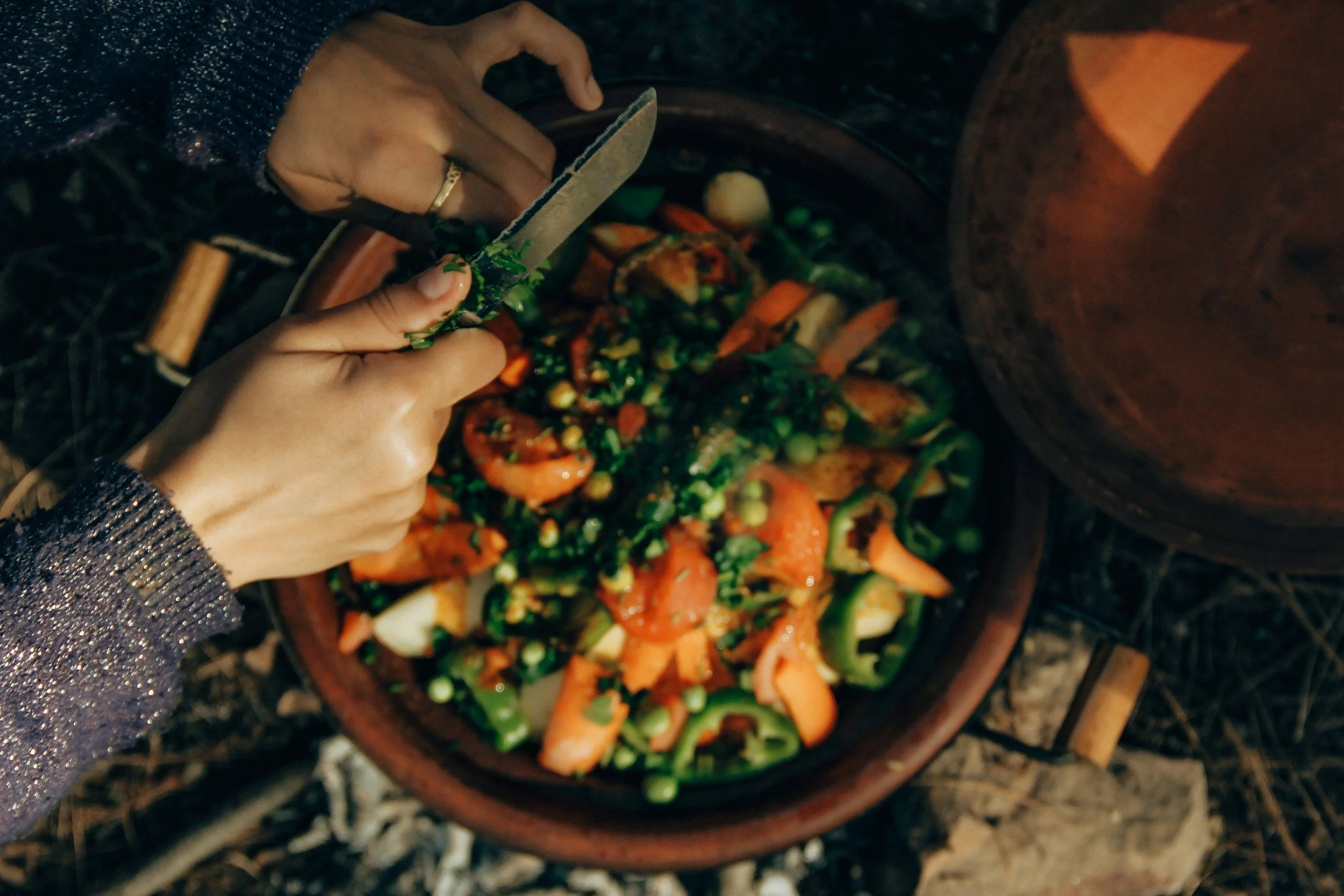 Person stirring a mixed vegetable dish in a clay pot over an open fire outdoors at night.