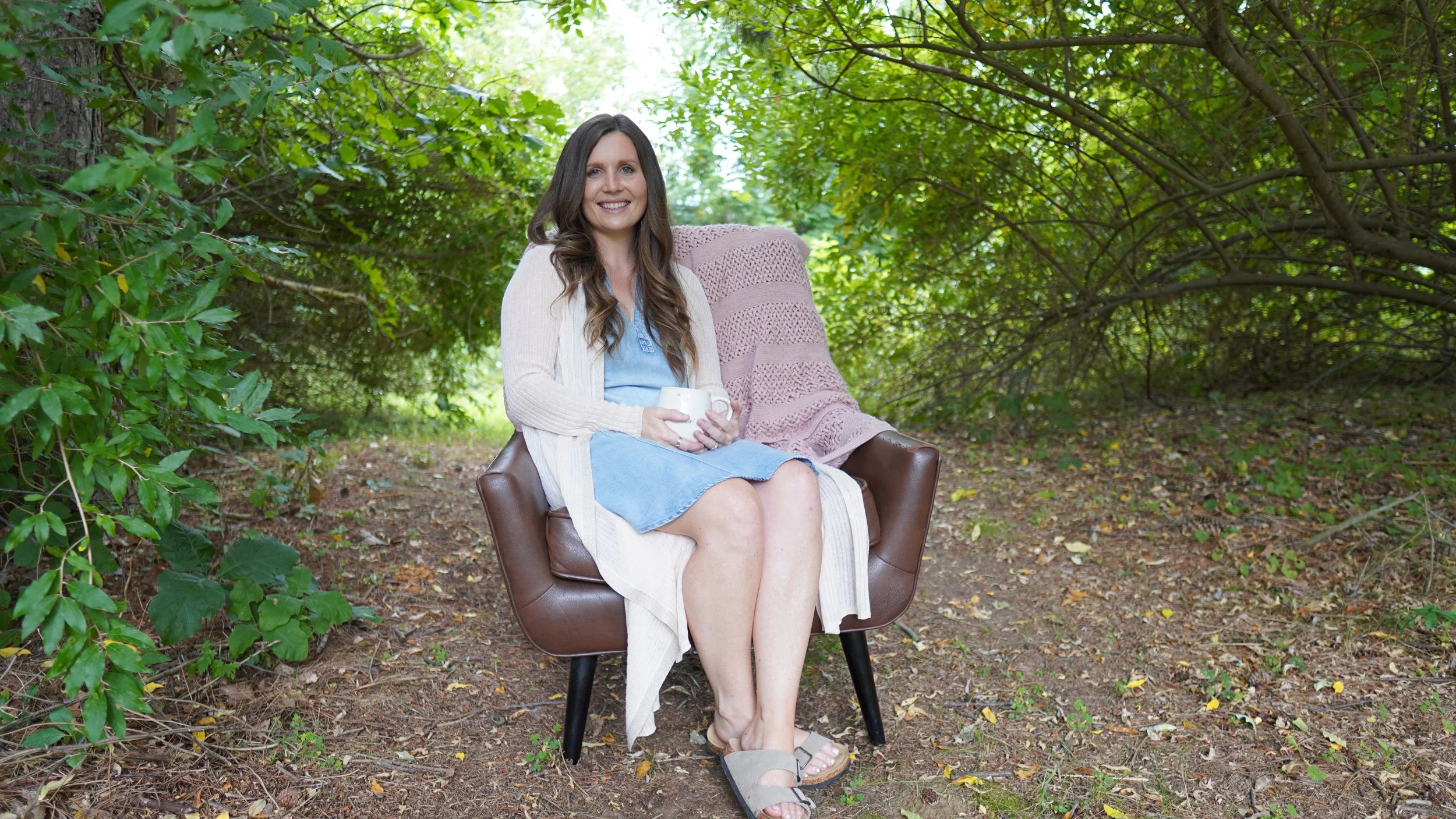A woman with long brown hair sitting on a brown chair outdoors in a wooded area, holding a white mug, smiling at the camera.