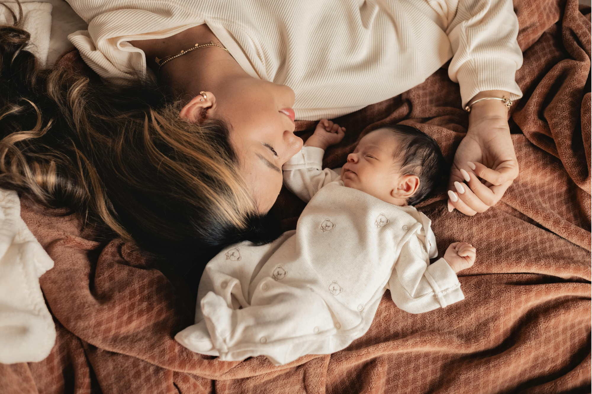 A woman lying down on a bed with a baby, both with their eyes closed, holding hands, surrounded by brown blankets.