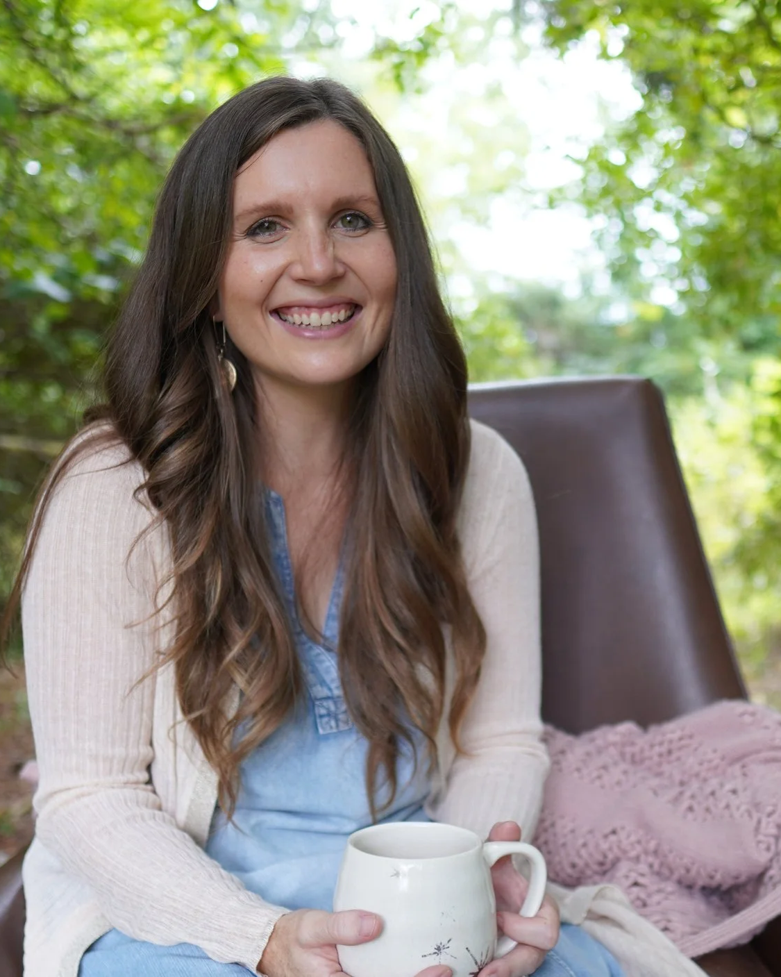 A woman with long brown hair smiling while sitting outdoors in a green, leafy setting, holding a white mug.