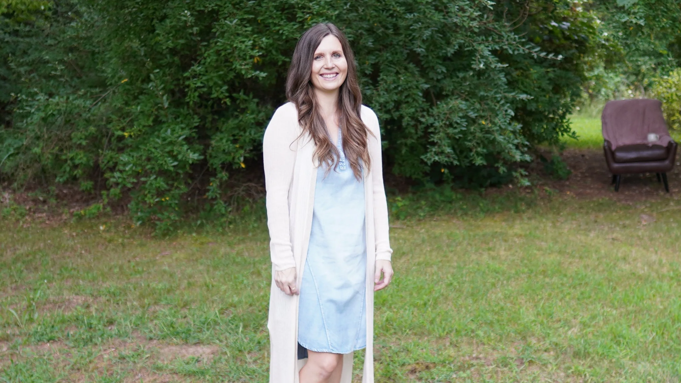 A woman with long brown hair wearing a white cardigan over a light blue dress standing outdoors on grass with trees and bushes in the background.