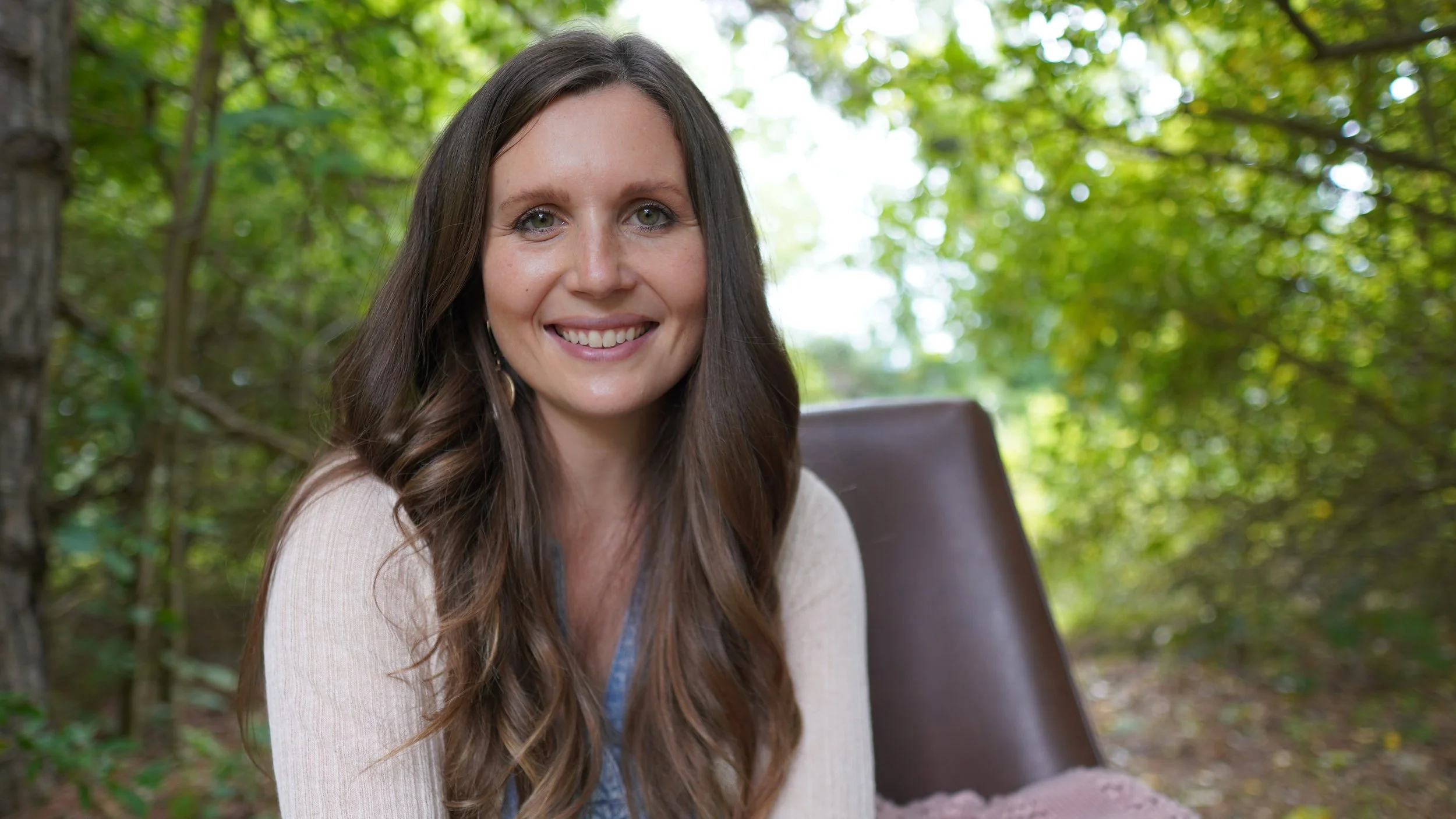 A smiling woman with long brown hair sitting outdoors in a wooded area with green leaves, on a wooden chair.