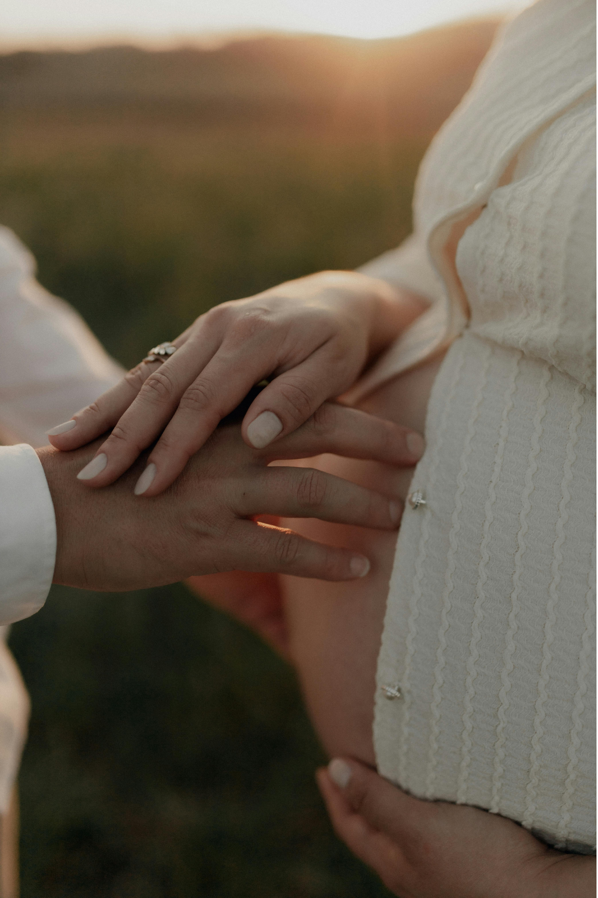Close-up of a couple holding hands during a sunset, with one person's hand resting on the other's chest, outdoors with a blurred background of hills or field.