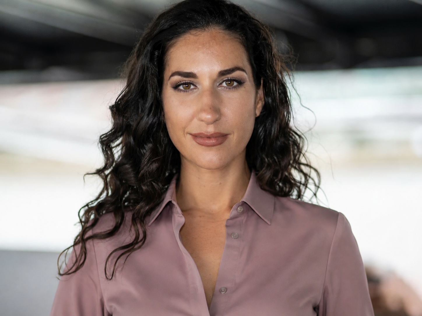 A woman with wavy dark hair and brown eyes wearing a light pink collared shirt, looking at the camera.