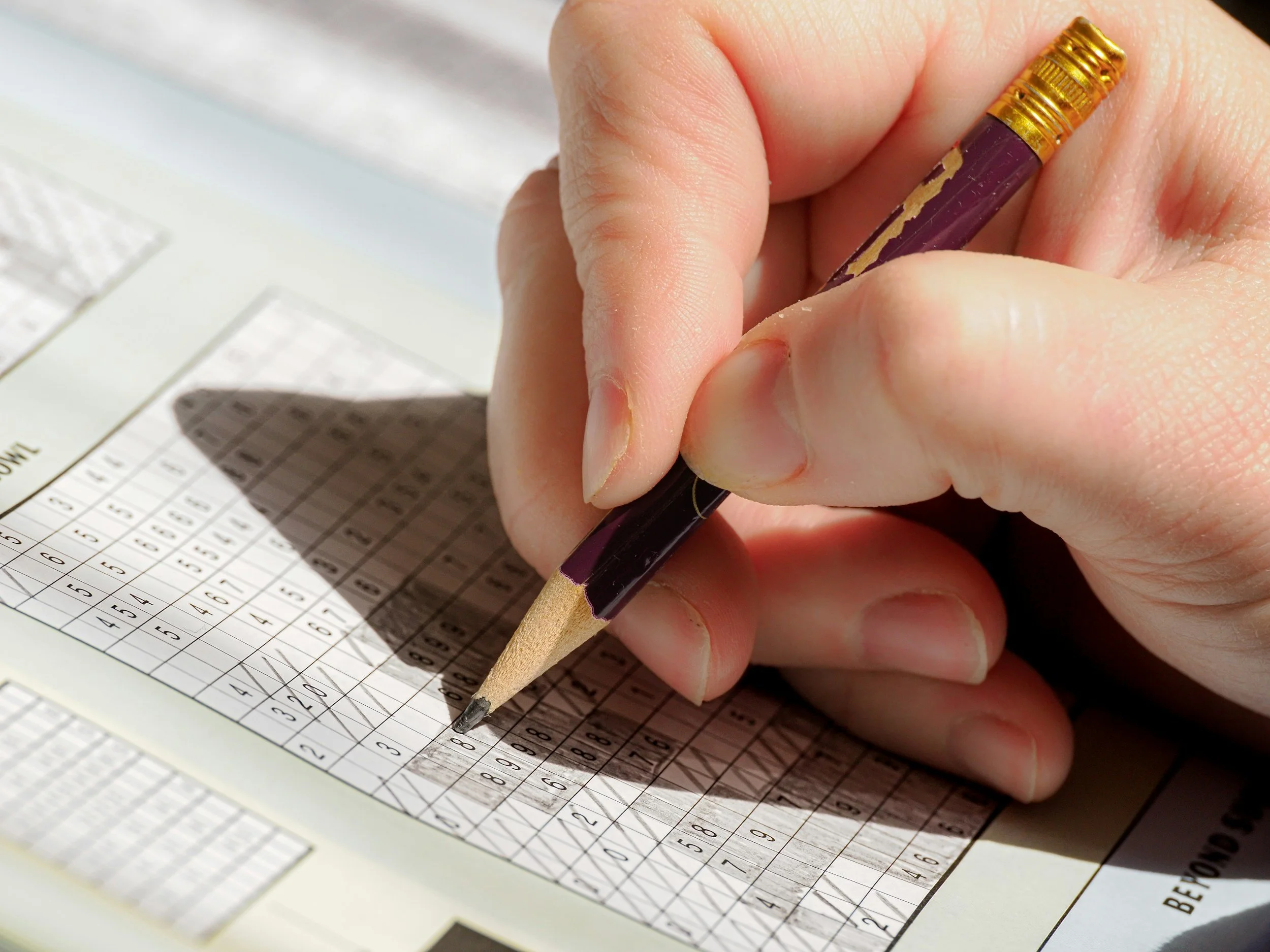 Close-up of a person's hand holding a pencil and filling out a bubble sheet answer sheet with a grid of numbers and multiple-choice options.