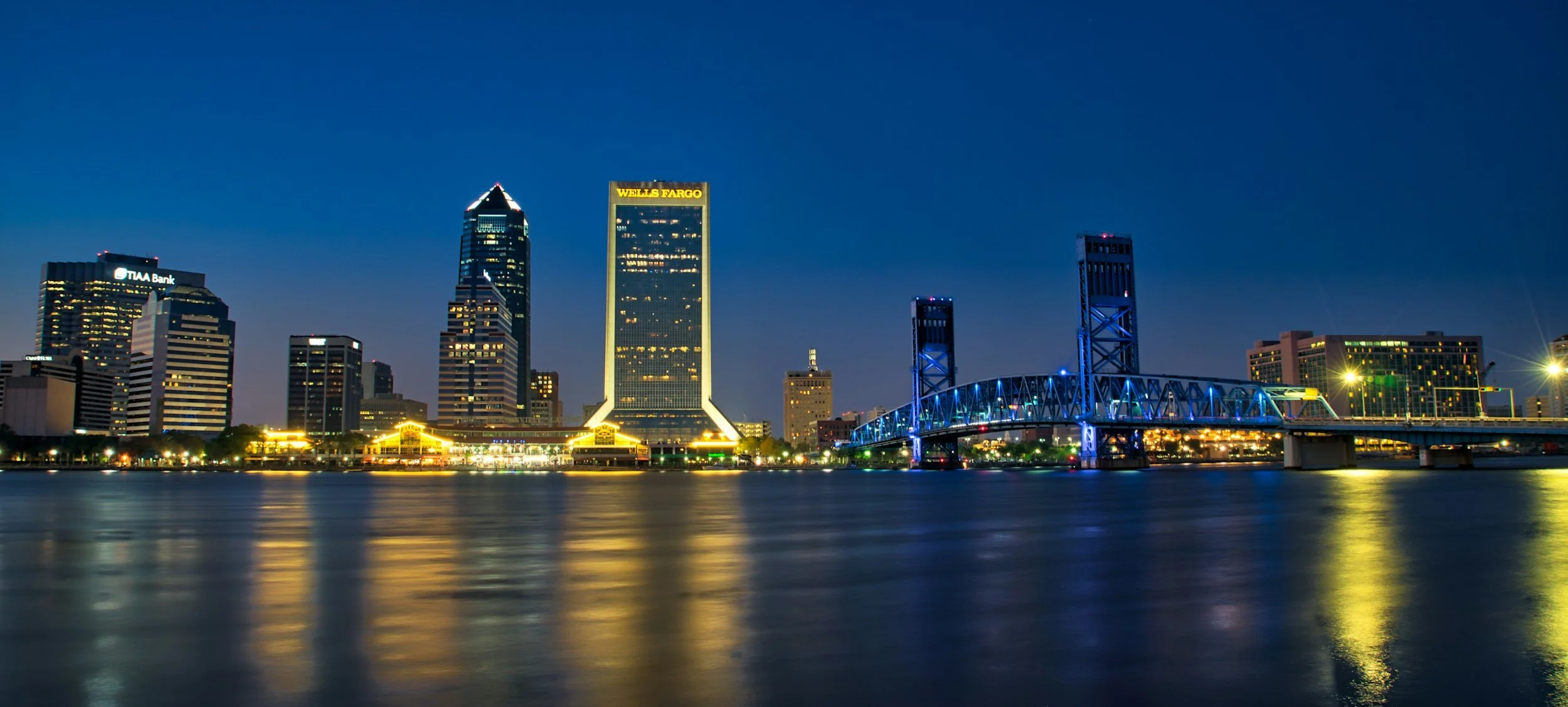 Nighttime view of a city skyline with illuminated buildings and a bridge over water, reflecting city lights.