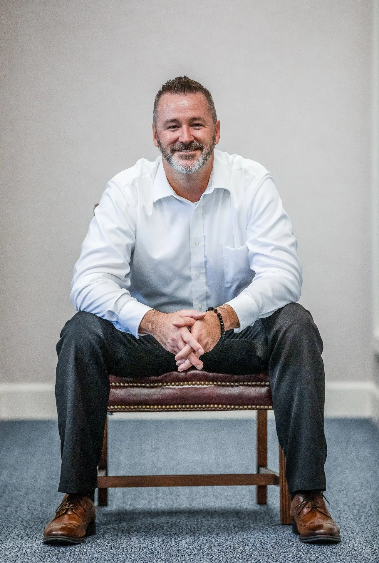 A middle-aged man with a beard and short hair, smiling, sitting on a chair with his hands clasped, wearing a white dress shirt, dark pants, and brown shoes, in a plain room with gray walls and blue carpet.