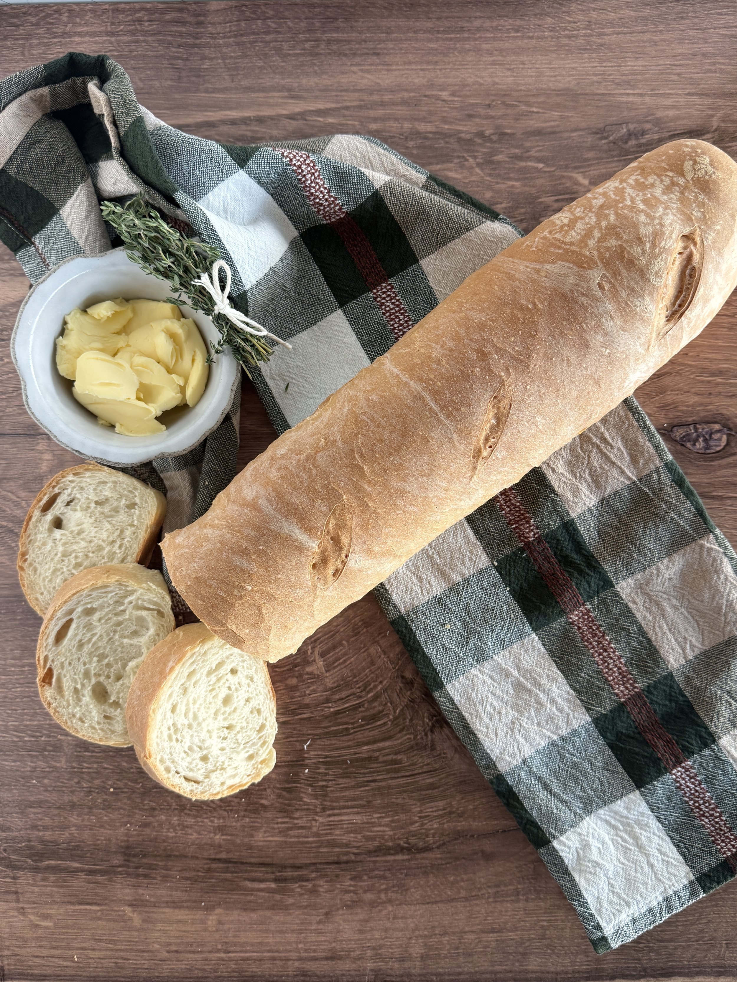 A large loaf of french bread with three slices sliced from it beside a dish of butter and thyme.