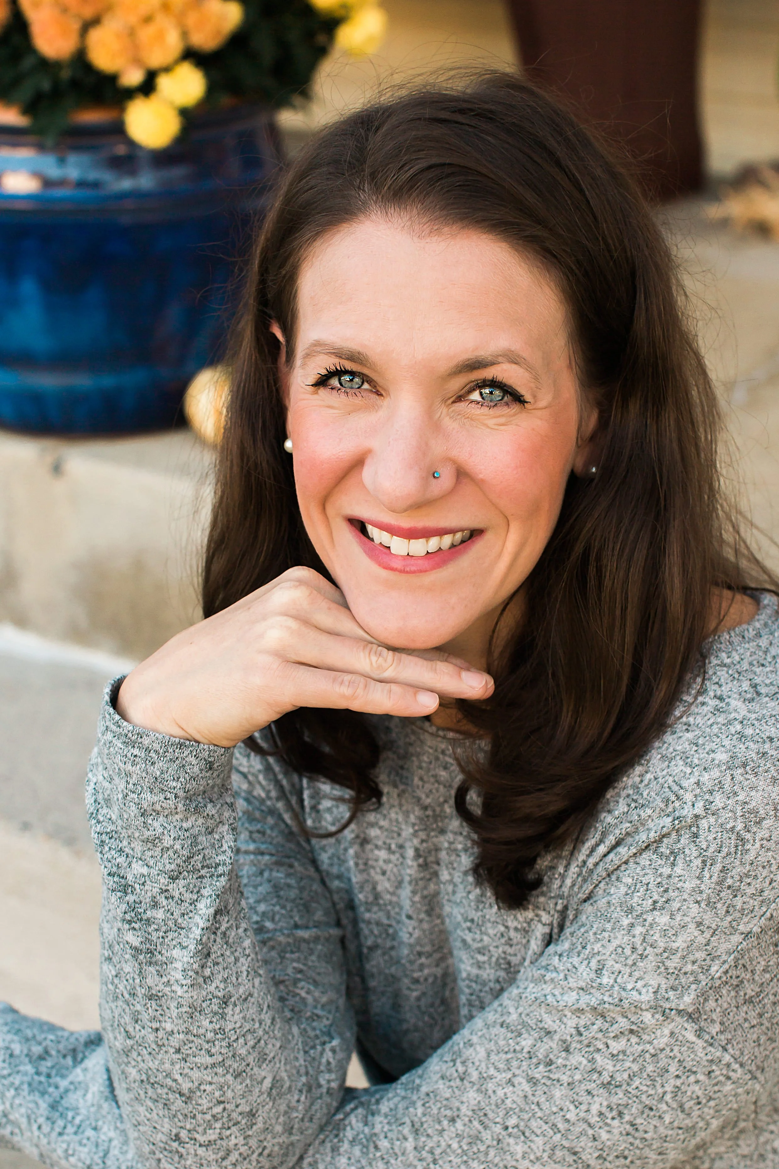 A woman with long dark hair, blue eyes, and a nose piercing, smiling and resting her chin on her hand, wearing a gray sweater, with yellow flowers in a dark blue vase in the background.
