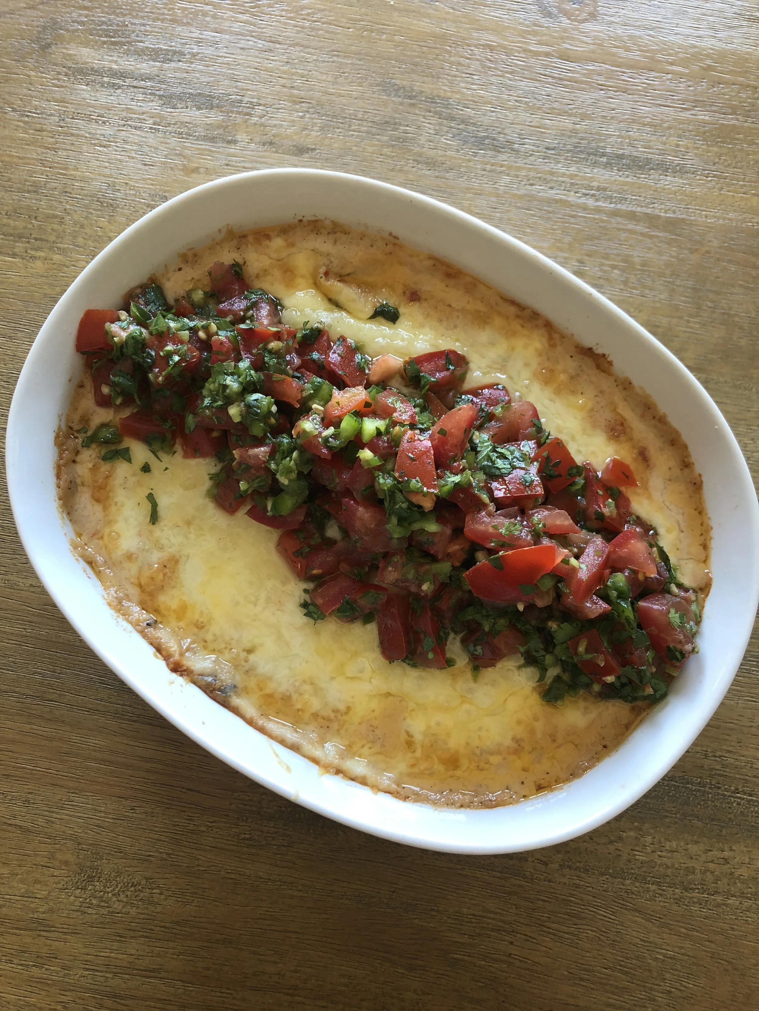 Baked dish with melted cheese, topped with chopped tomatoes, green onions, and herbs in a white oval dish on a wooden table.