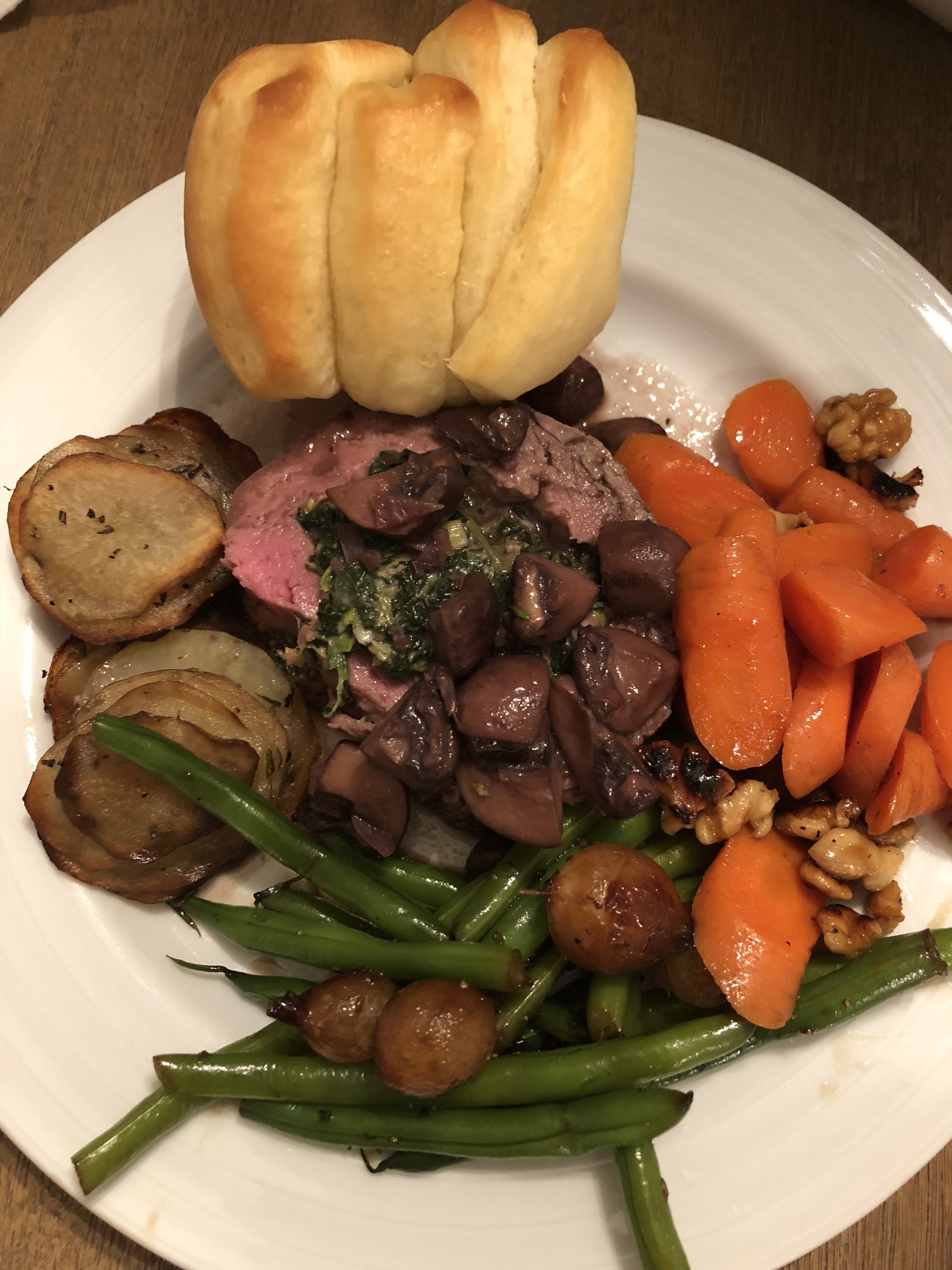 A white plate with a fresh roll, roasted potatoes, cooked carrots, sautéed green beans, and a serving of sliced beef with herbs and sauce.
