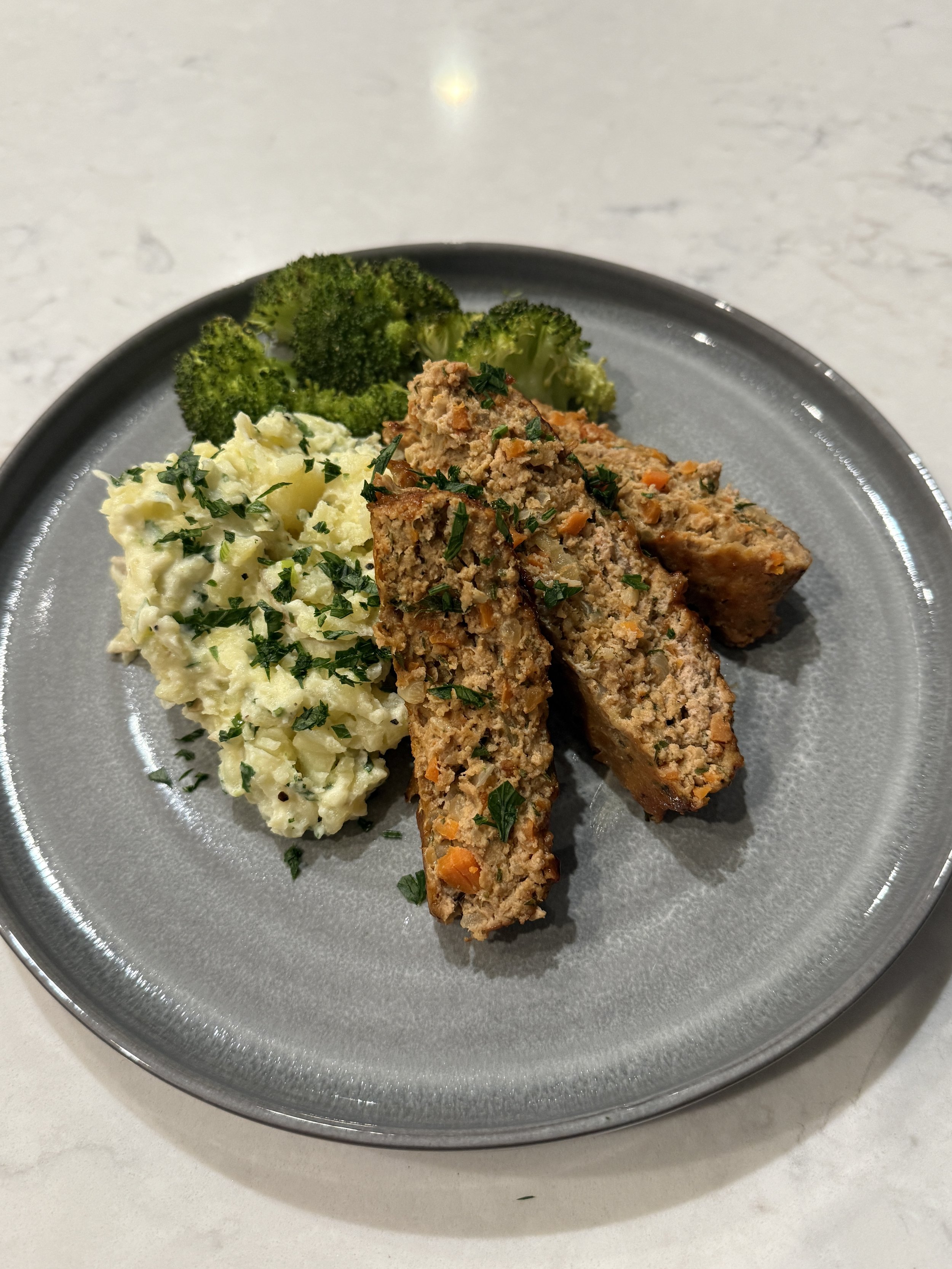 A plate with mashed potatoes sprinkled with parsley, steamed broccoli, and two slices of meatloaf garnished with parsley.
