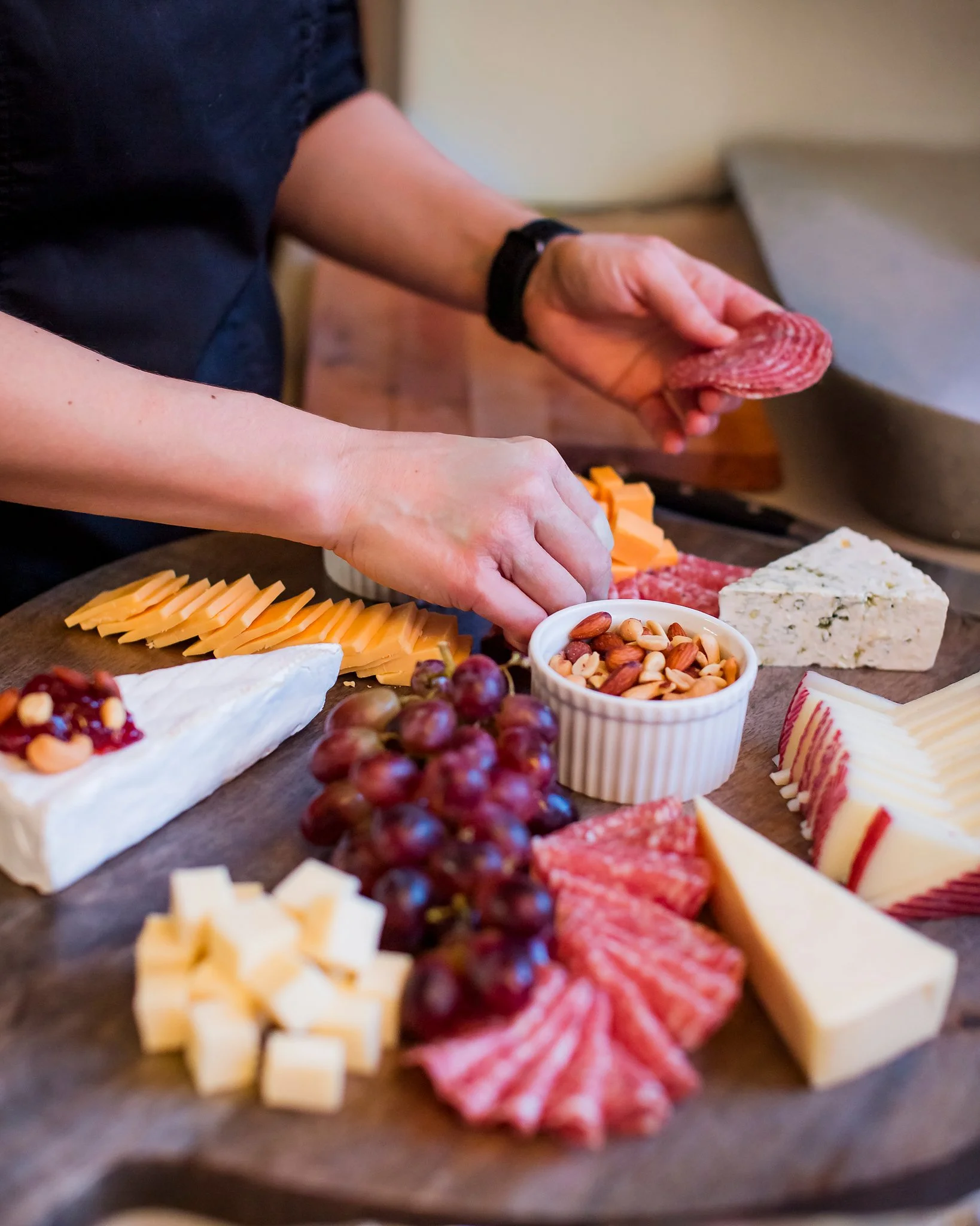 A charcuterie board with assorted cheeses, sliced meats, grapes, nuts, and a small dish of dried fruit or nuts.