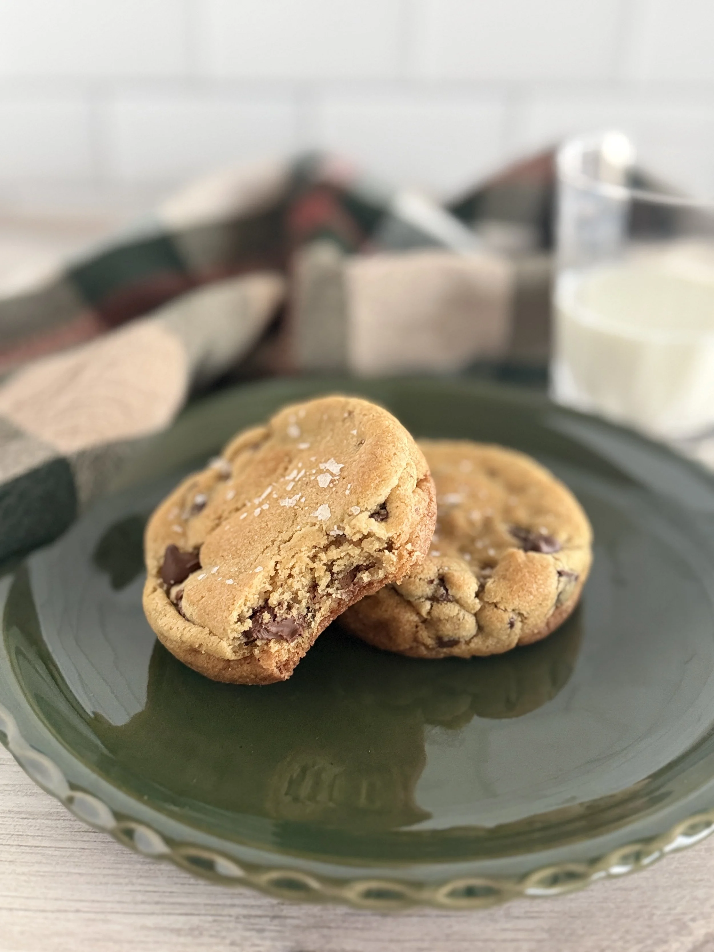 Two golden chocolate chip cookies with flakly sea salt on a dark green plate.