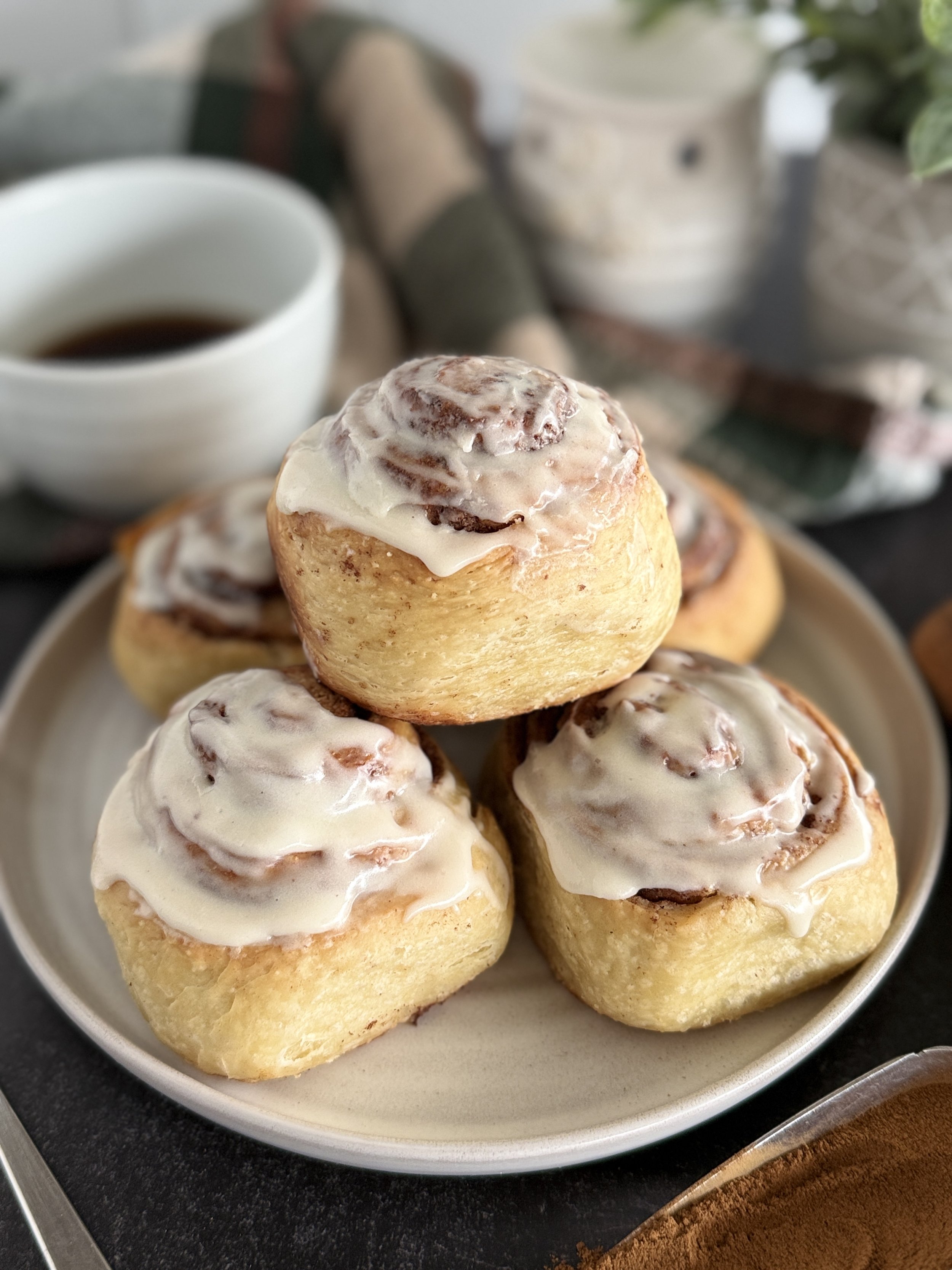 Close up of five cinnamon rolls on a white plate with coffee in the background.