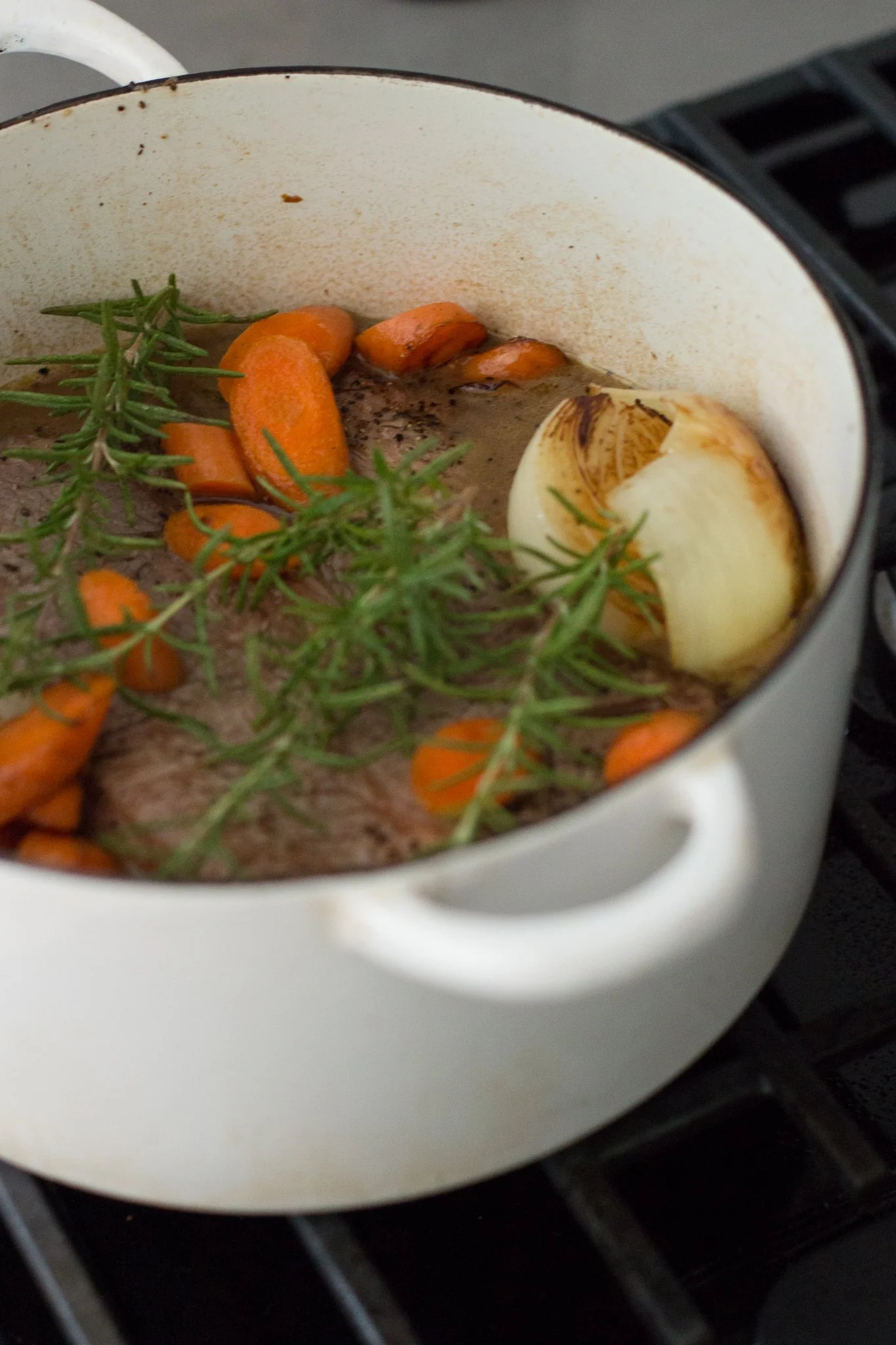A white enamel pot on a stove with whole garlic, carrots, and sprigs of rosemary inside, cooking in liquid.