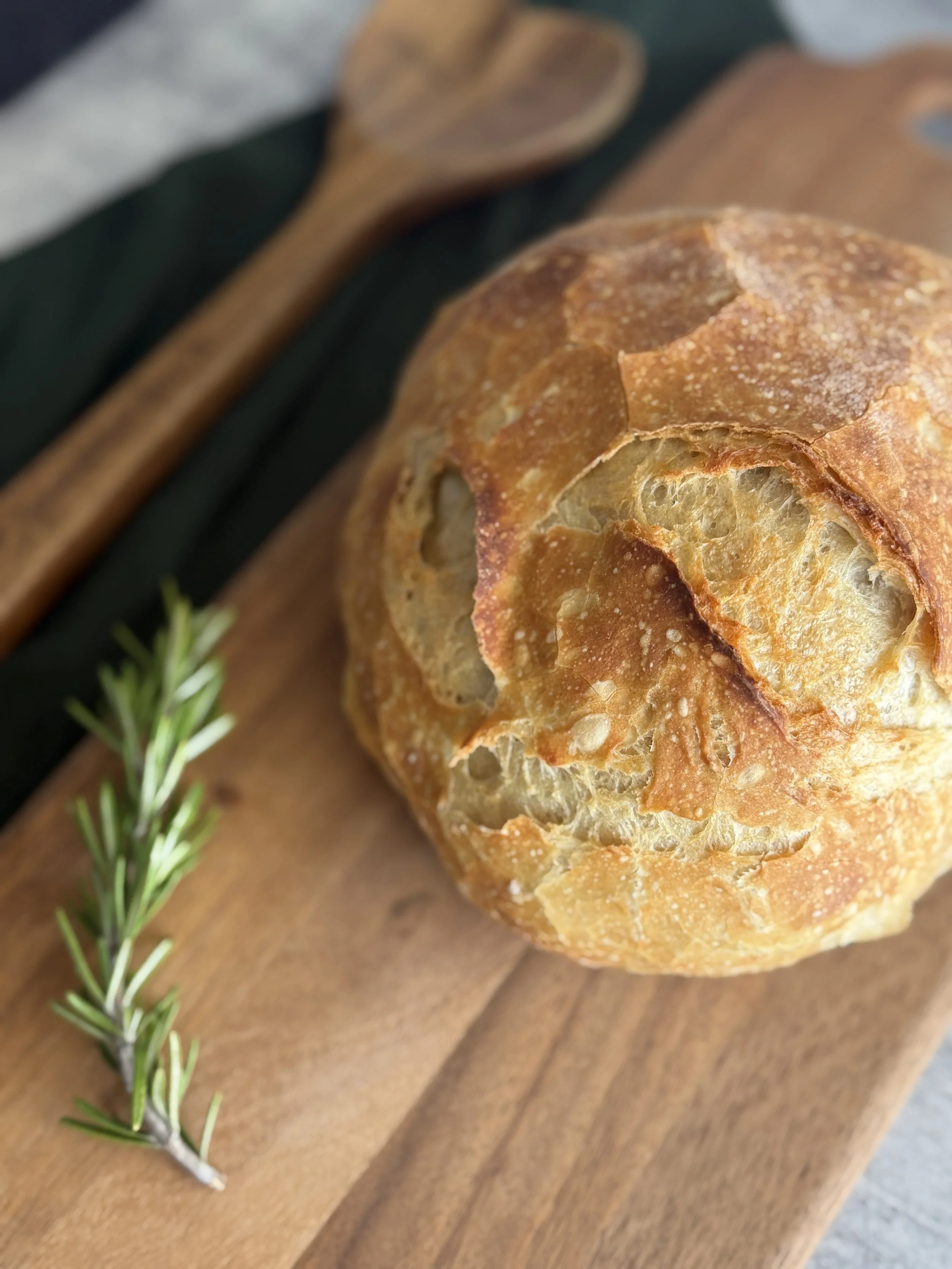 A loaf of crusty bread placed on a wooden cutting board with a sprig of rosemary and a wooden spoon nearby.