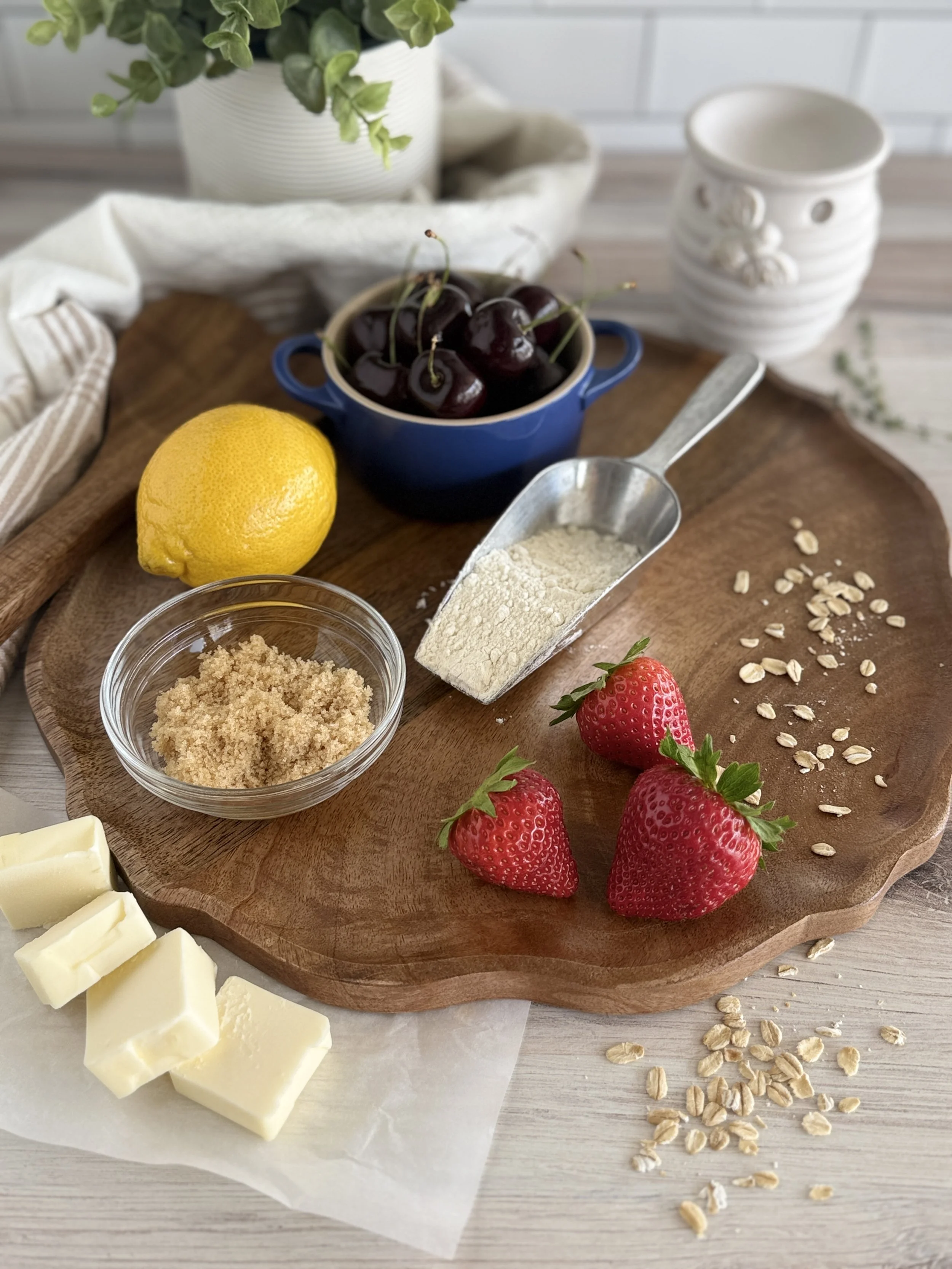 Fresh strawberries, cherries in a small blue bowl, a lemon, white chocolate, oats, and baking ingredients on a wooden cutting board with a potted plant in the background.