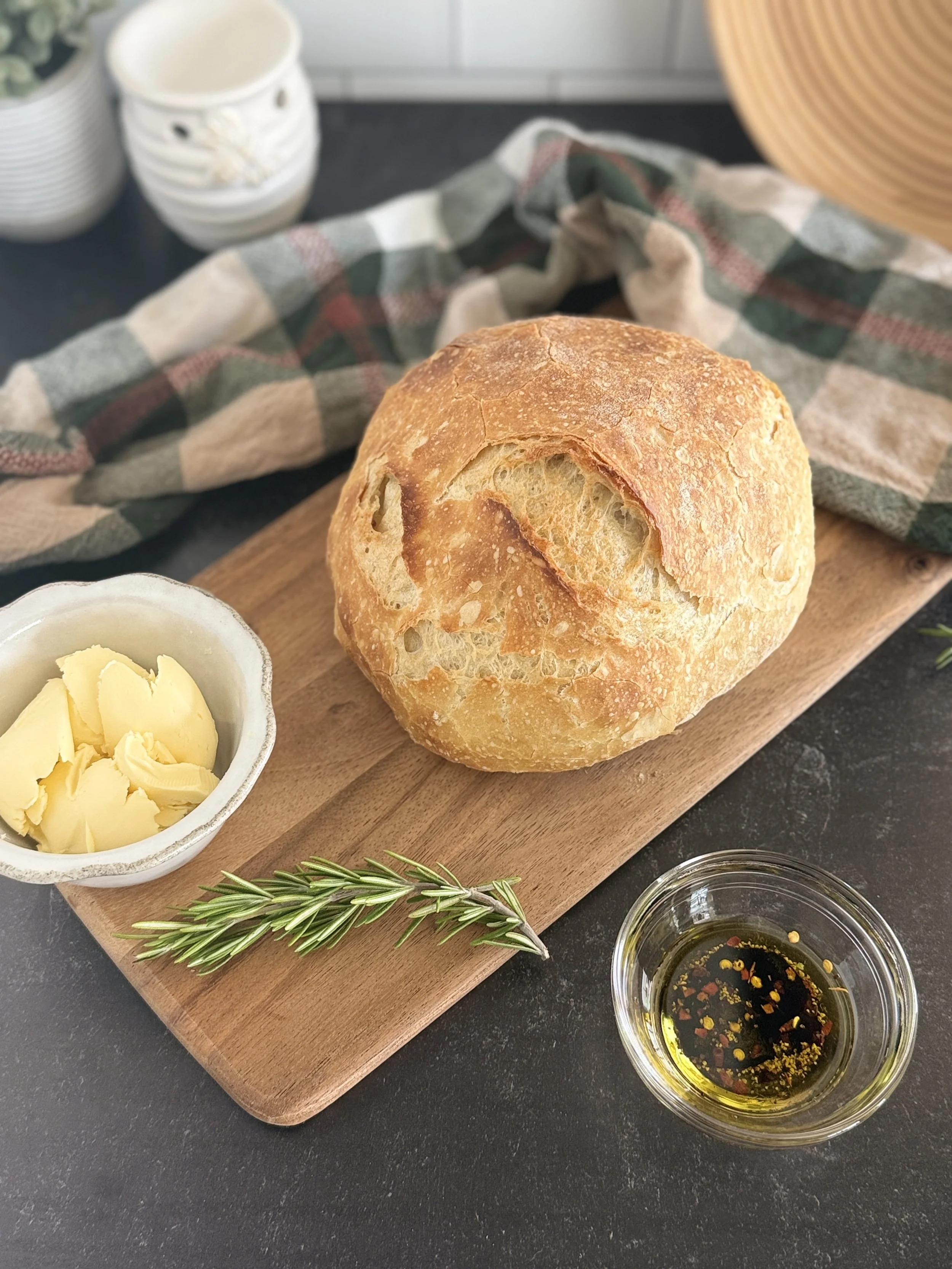 A loaf of crusty bread on a board with a bowl of butter beside it and rosemary.