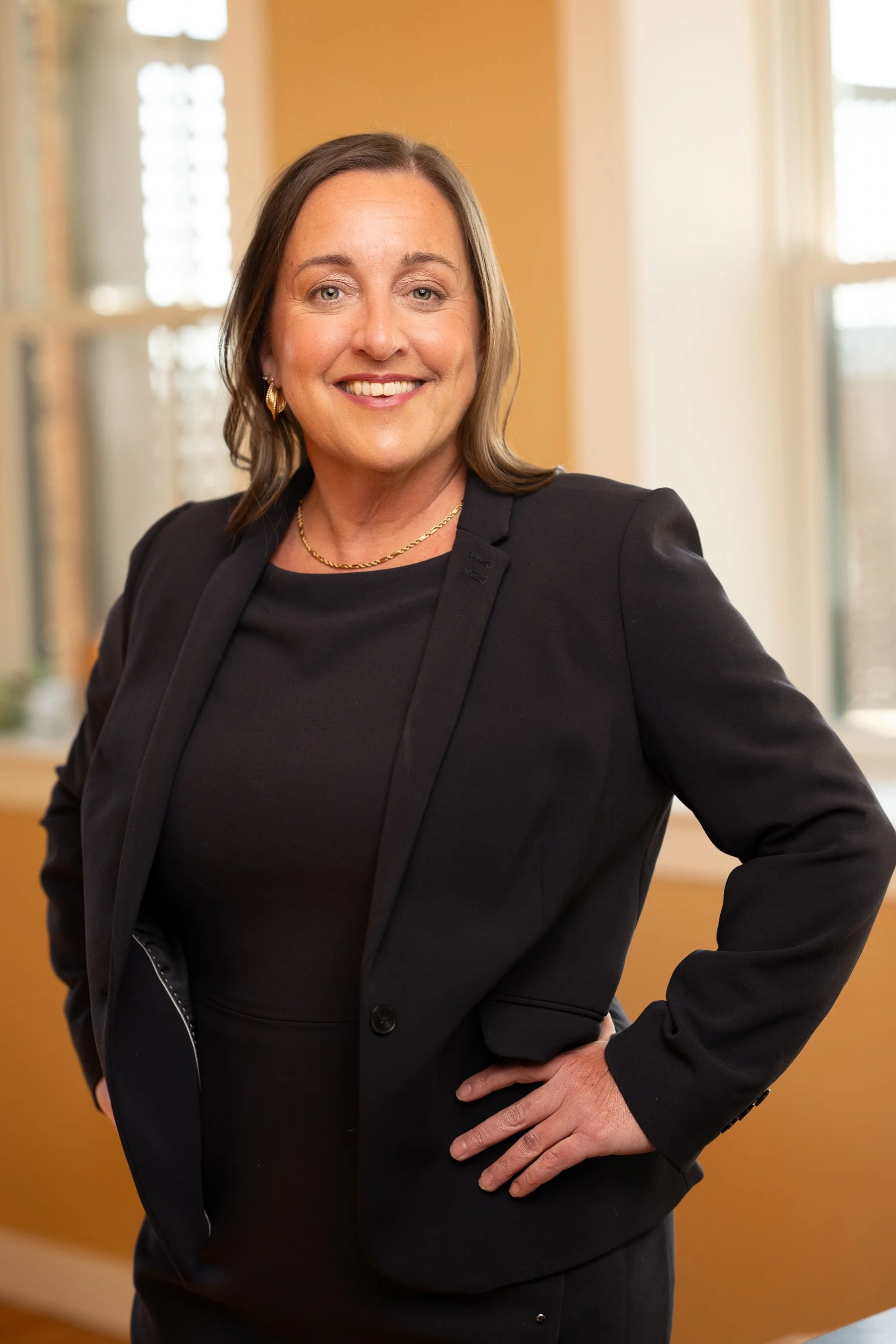 Gloucester attorney Christina Griecci of Griecci Law, standing indoors with a warm-colored wall and windows in the background, smiling and looking at the camera.