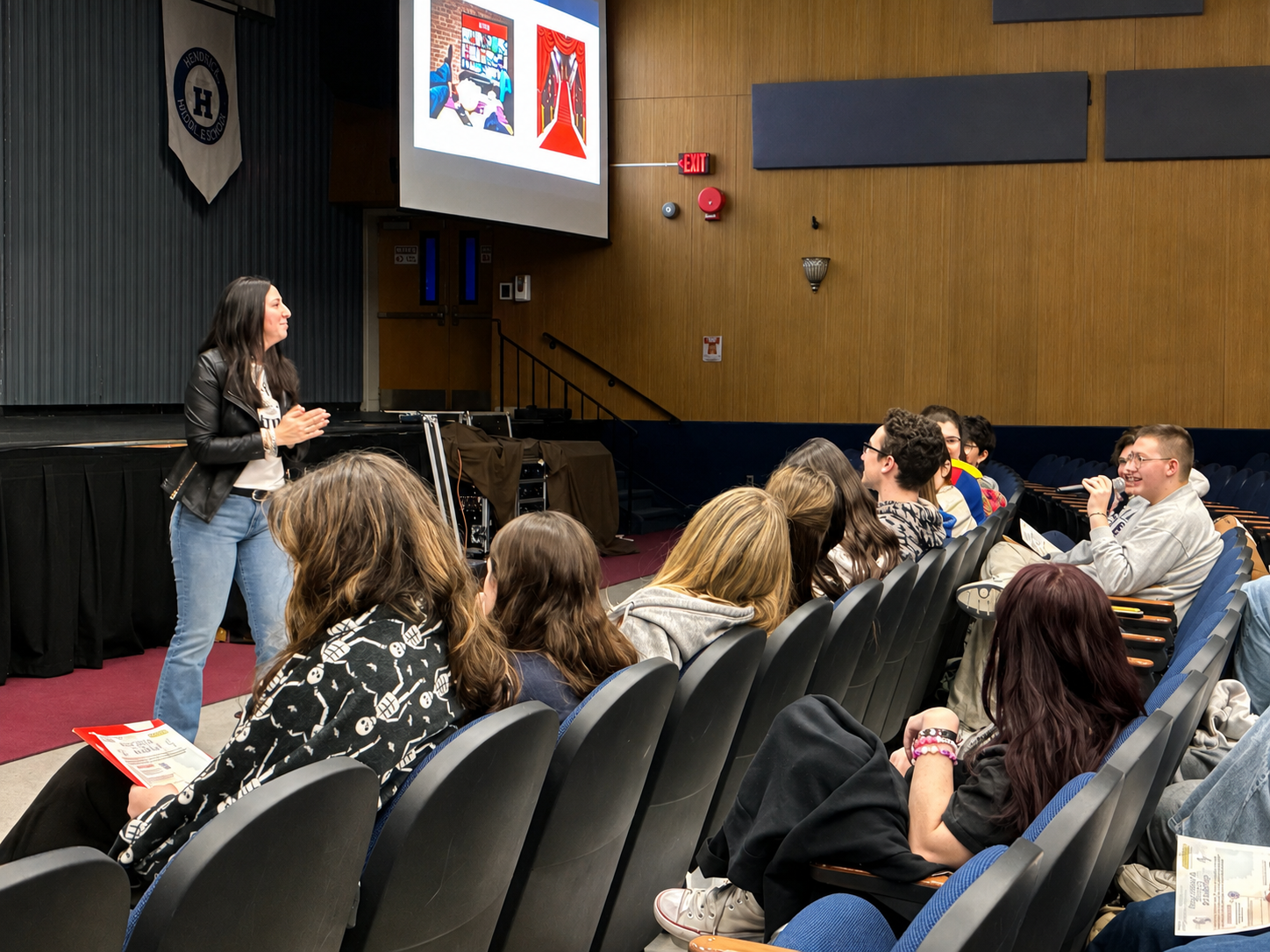 A woman in a leather jacket speaking to an audience of students in a lecture hall, with some students holding papers and one student holding a microphone.
