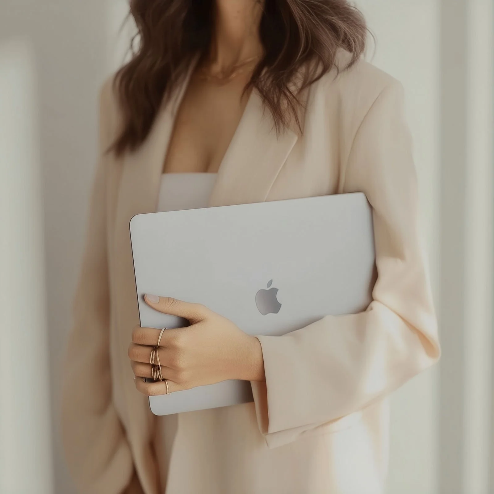 A woman wearing a beige blazer holding a silver MacBook laptop.