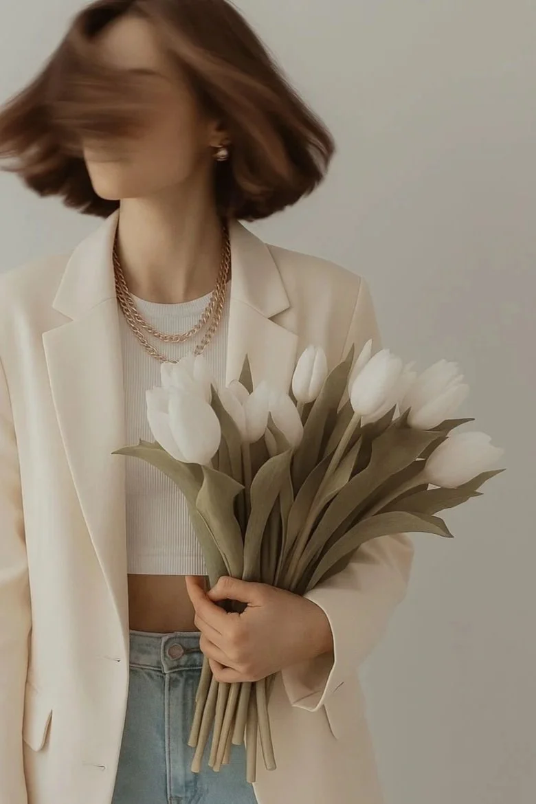 Person with shoulder-length brown hair holding a bouquet of white tulips, wearing a beige blazer, white crop top, and layered gold chains.