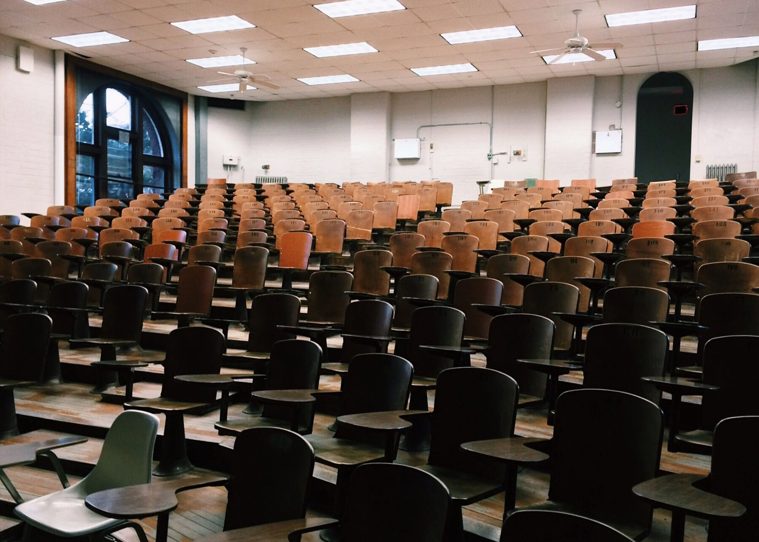 Empty classroom with wooden desks and black chairs arranged in rows, large arched window, white brick walls, ceiling fans, ceiling lights, and some wall-mounted equipment.