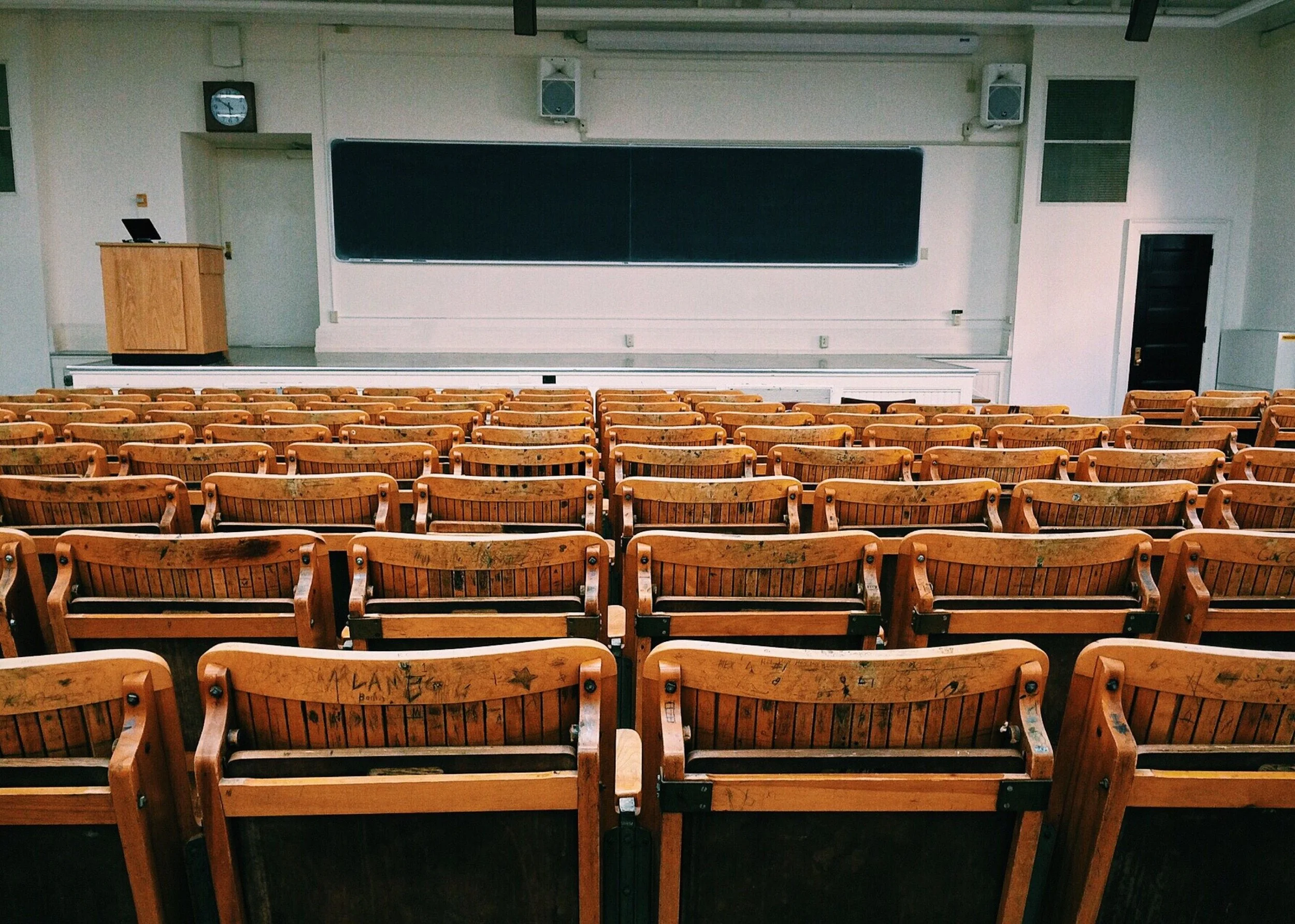Empty classroom with rows of wooden student desks, a blackboard, a wooden podium, and a clock on the wall.