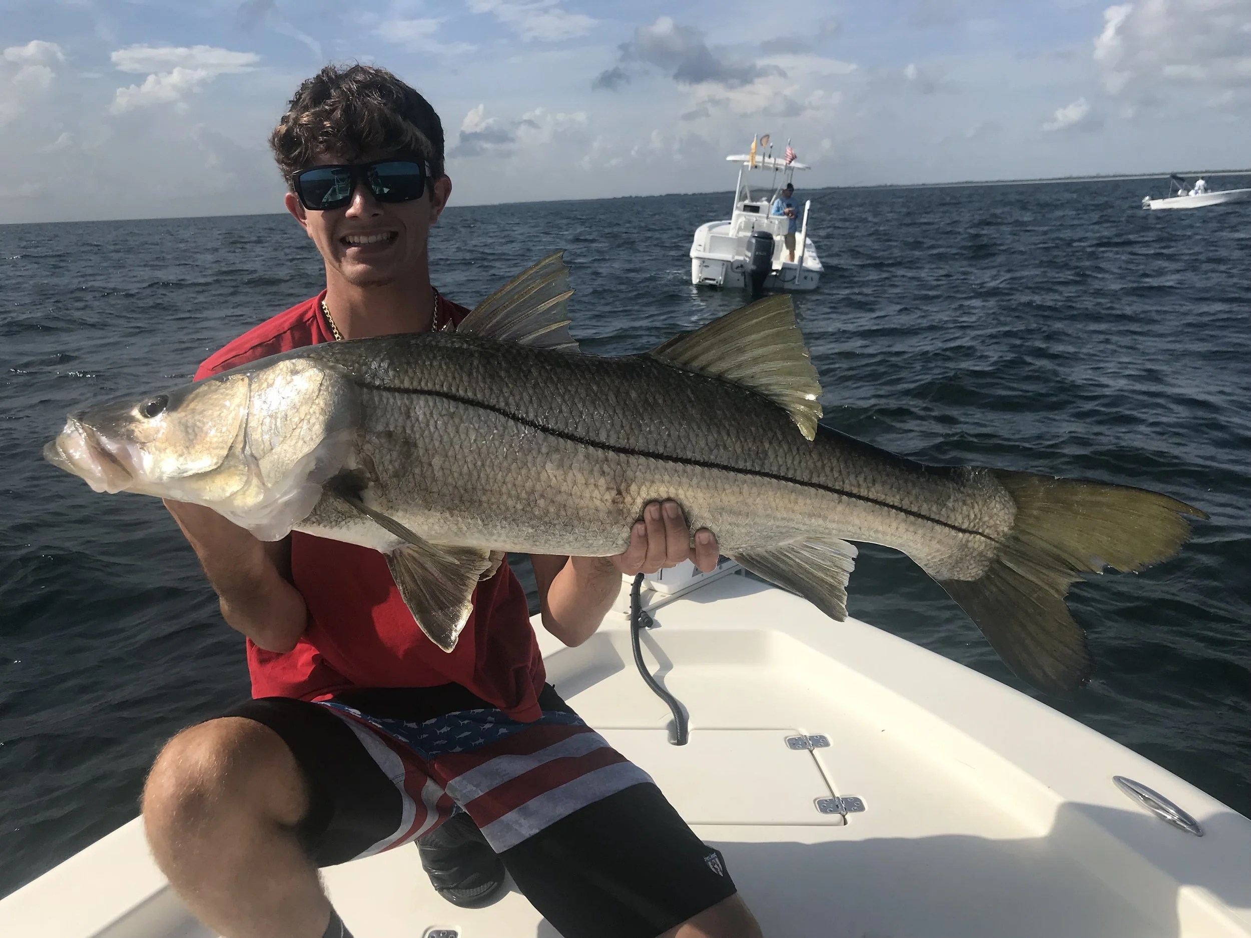 A man wearing sunglasses and a red shirt holding a large fish on a boat at sea during daytime.