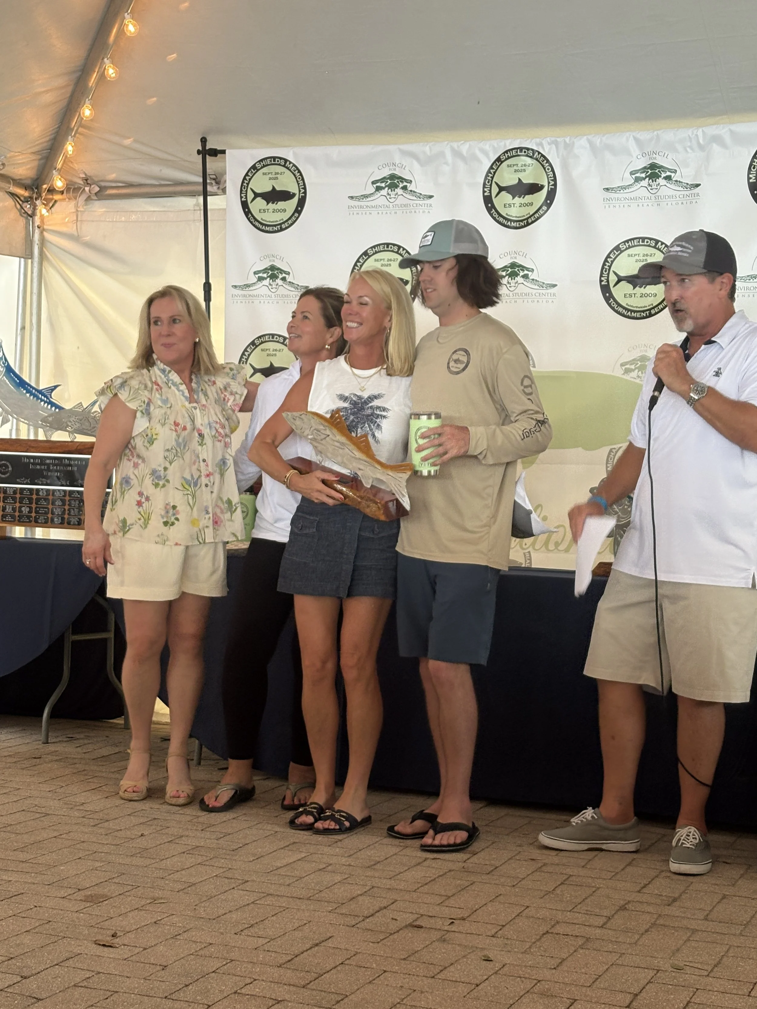 People standing under a tent during an award ceremony. Four women and two men, some holding a plaque or a drink, smiling. A backdrop with logos related to environmental studies and a tournament series.