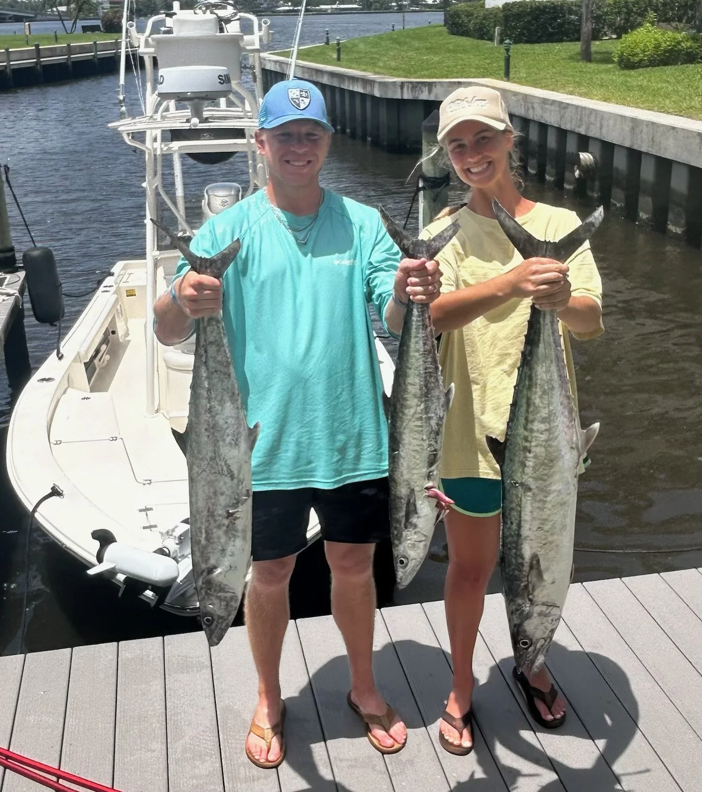Two people standing on a dock holding large fish they caught. The person on the left is wearing a blue cap, aqua shirt, black shorts, and flip-flops. The person on the right is wearing a beige cap, yellow shirt, green shorts, and flip-flops. A boat is docked behind them on a waterway, with greenery and a grassy area nearby.