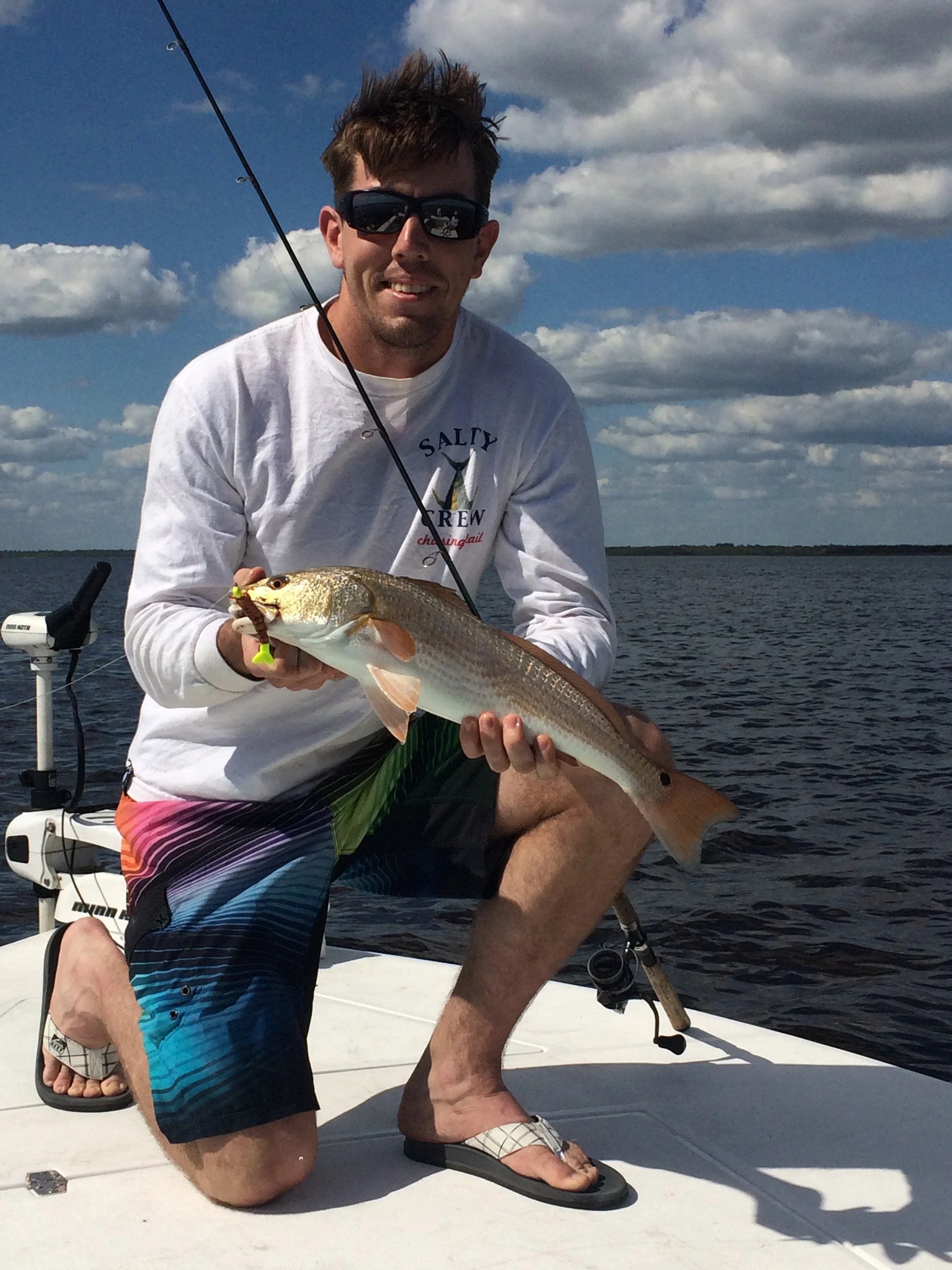 A man on a boat holding a fish he caught, wearing sunglasses, a white shirt, and colorful shorts, with a cloudy sky and water in the background.