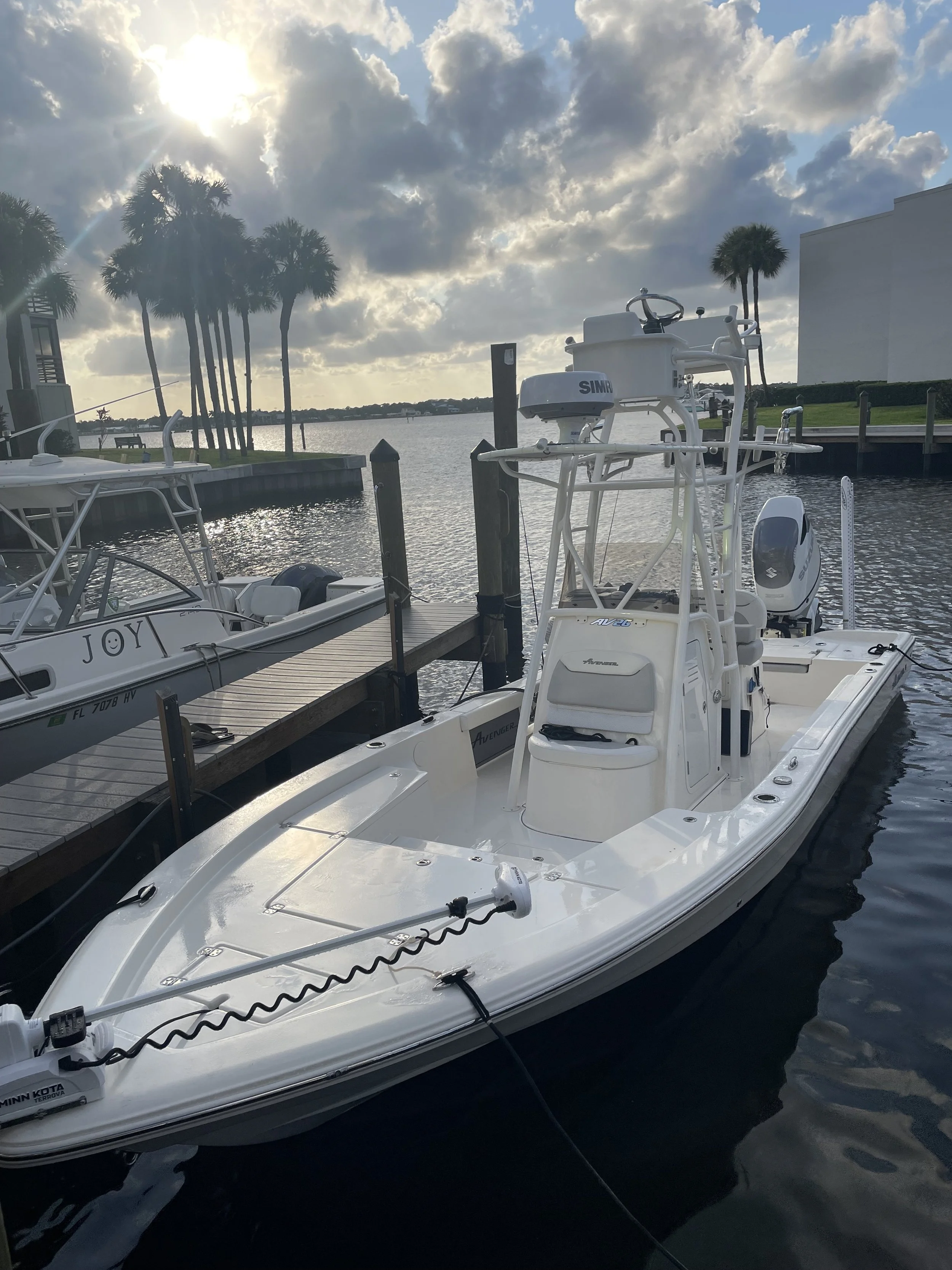 A white motorboat docked at a marina on a cloudy day, with palm trees and water in the background.